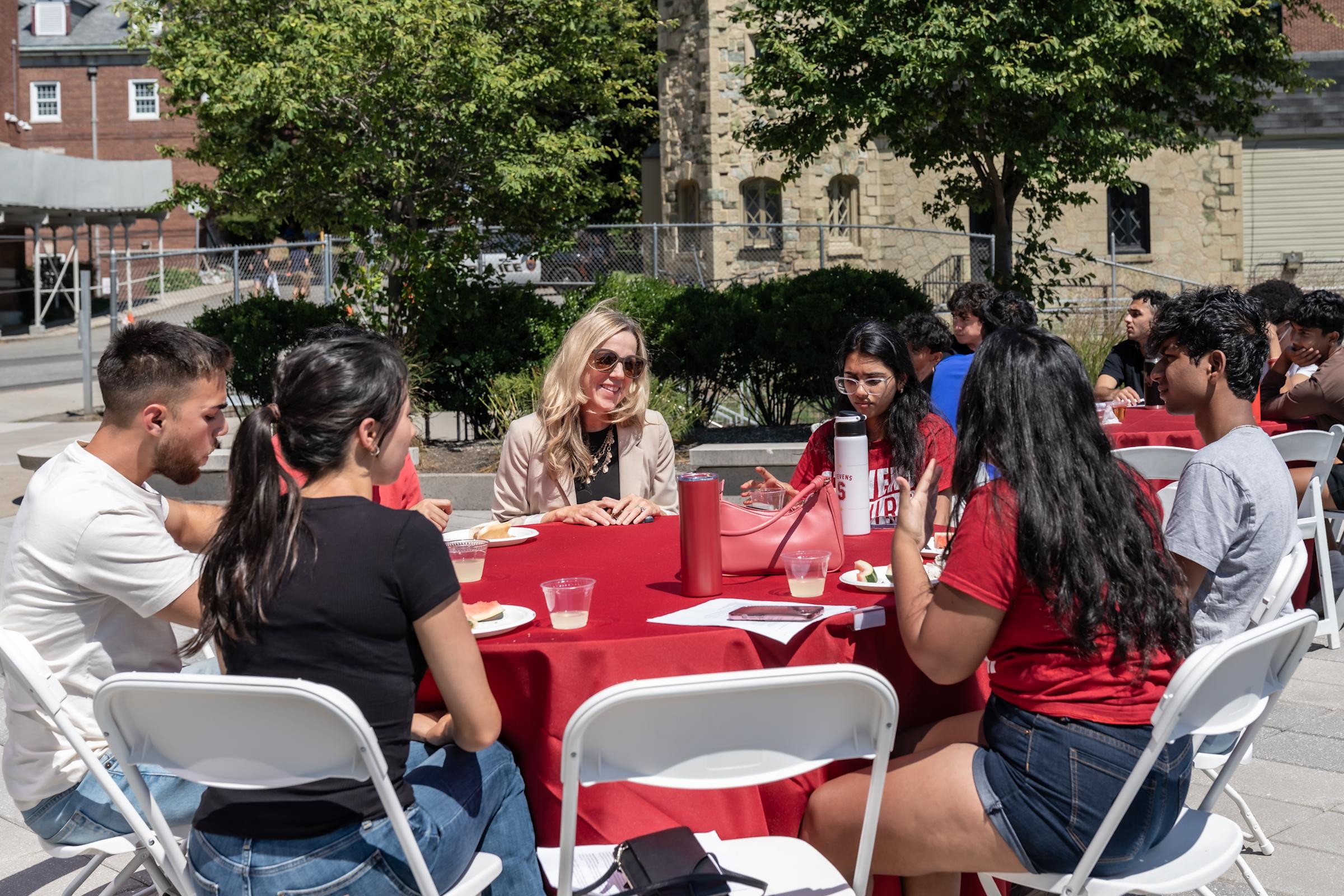 Blonde woman in beige blazer dining with diverse group of students at red-tablecloth outdoor meal on campus plaza with historic stone buildings.