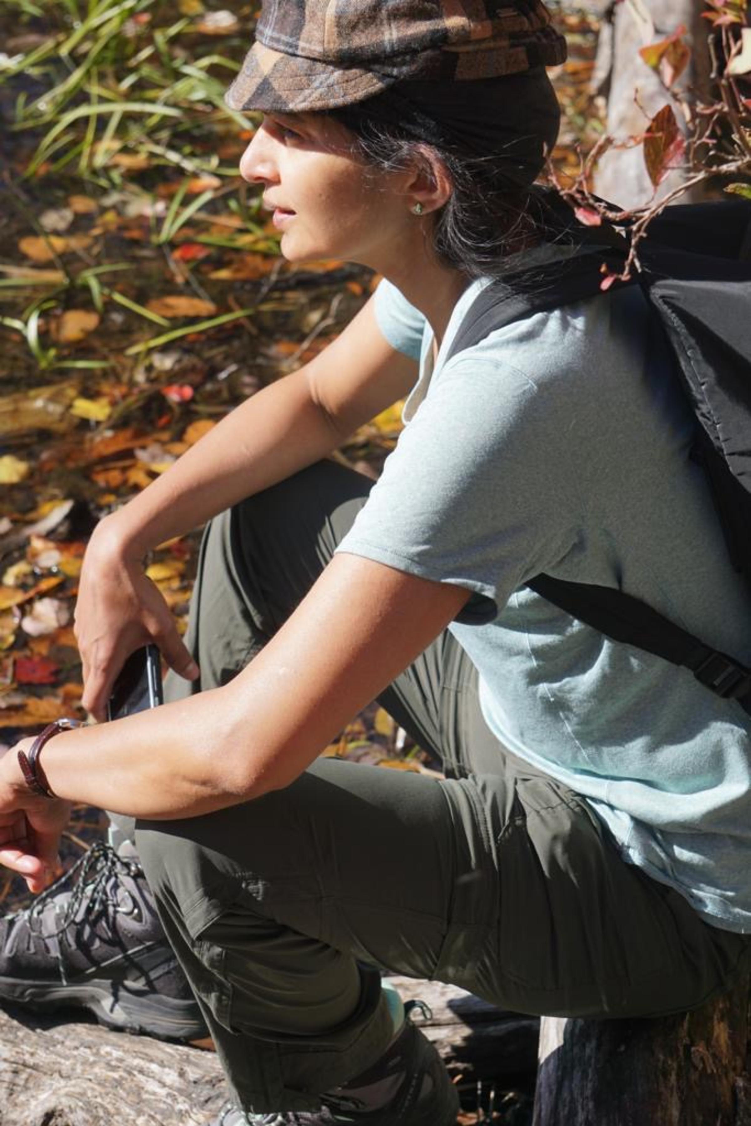 A woman in a blue T-shirt, gray pants, hiking boots and hat sits in a rock in a forested area.