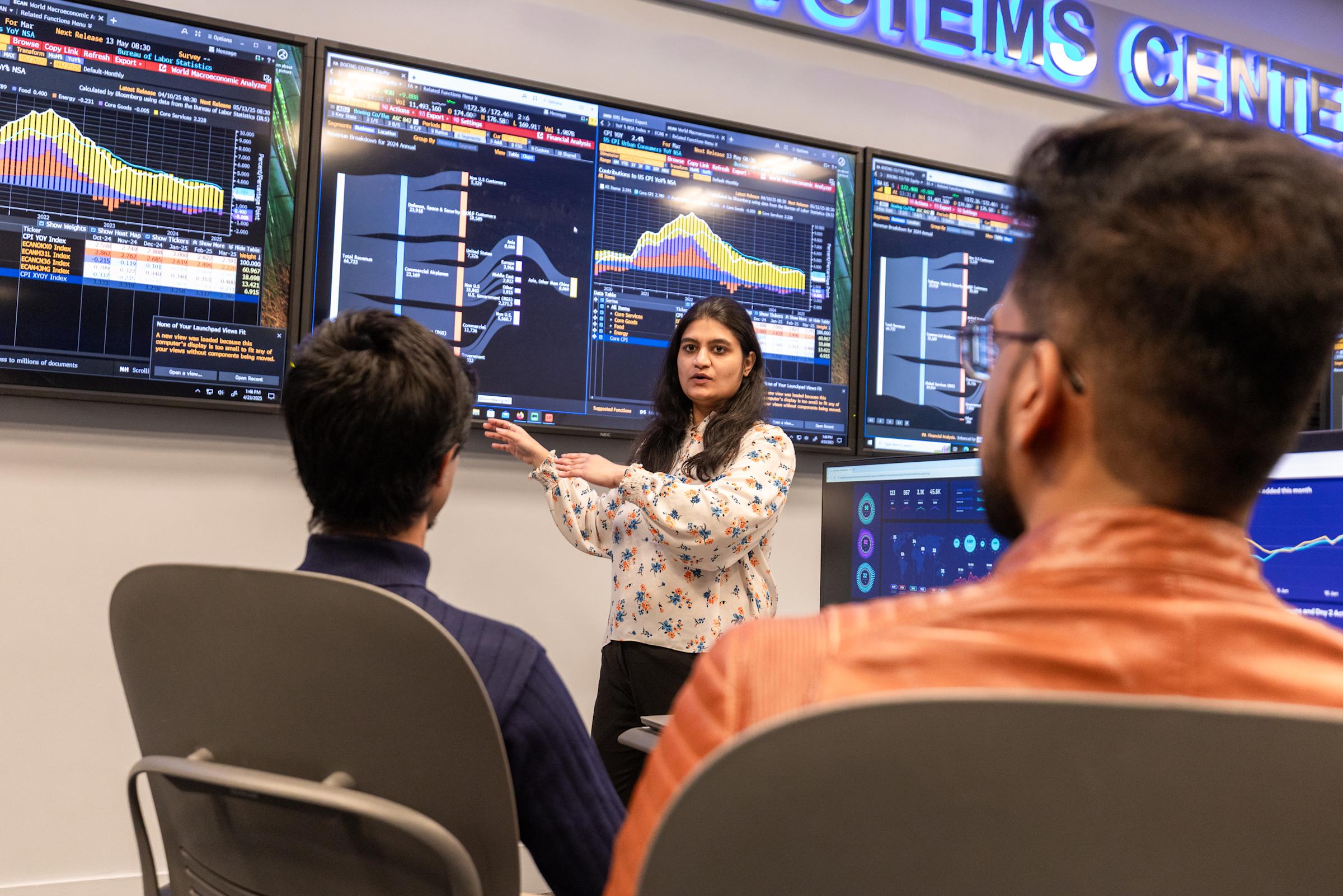 A female instructor in a floral blouse presents to students in front of a wall of large monitors displaying financial data, charts, and Bloomberg terminal screens in the Systems Center. Two male students sit facing her in the trading simulation room.