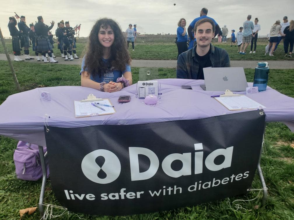 Arianna Gehan ’24 and Frank Pinnola ’23 sit outdoors at a table with a banner that reads "Daia - live safer with diabetes"