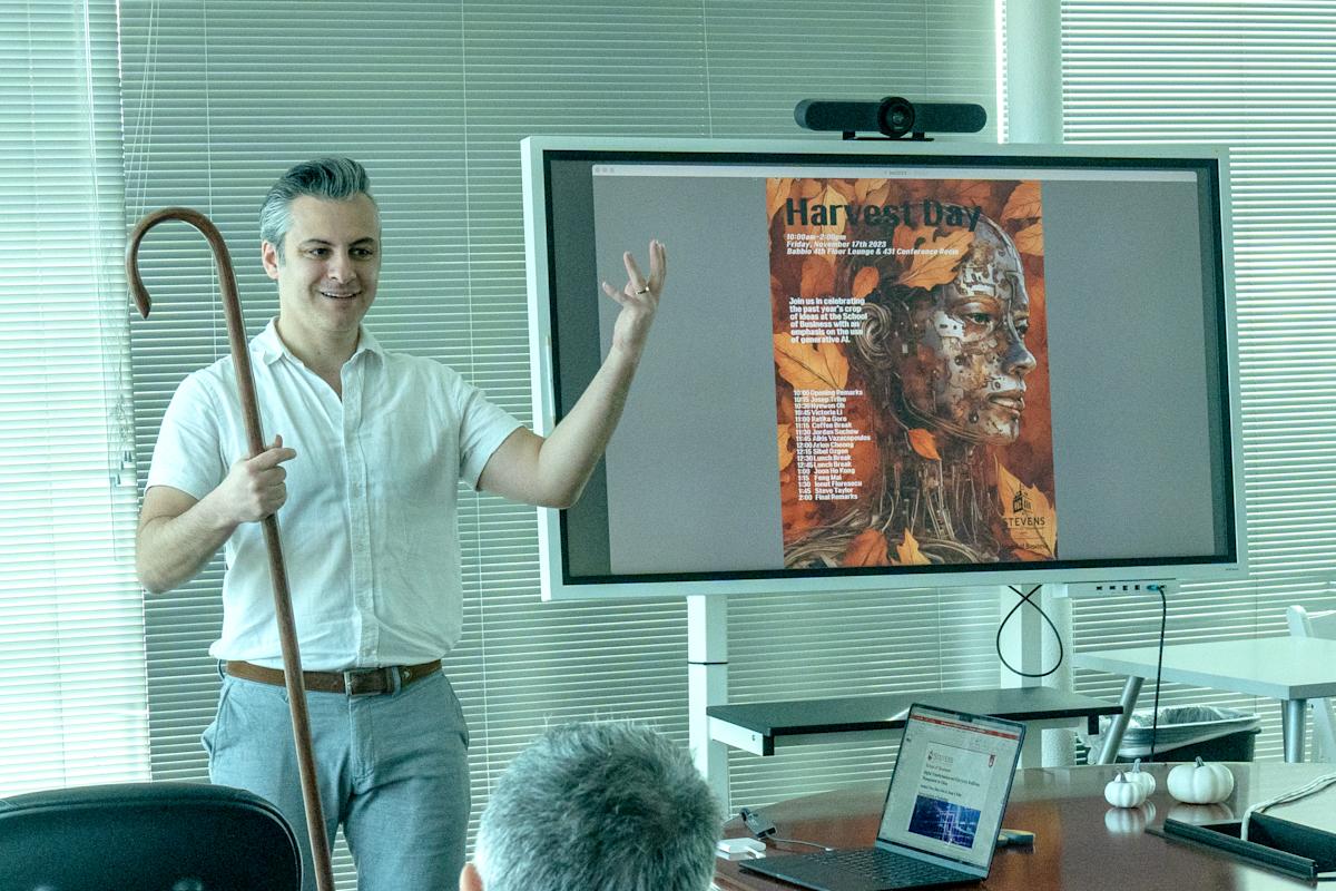Jordan Suchow holds a shepherd's crook next to a smart board with an AI generated photo of a robot on a background of autumn leaves with the Harvest Day schedule on top. 
