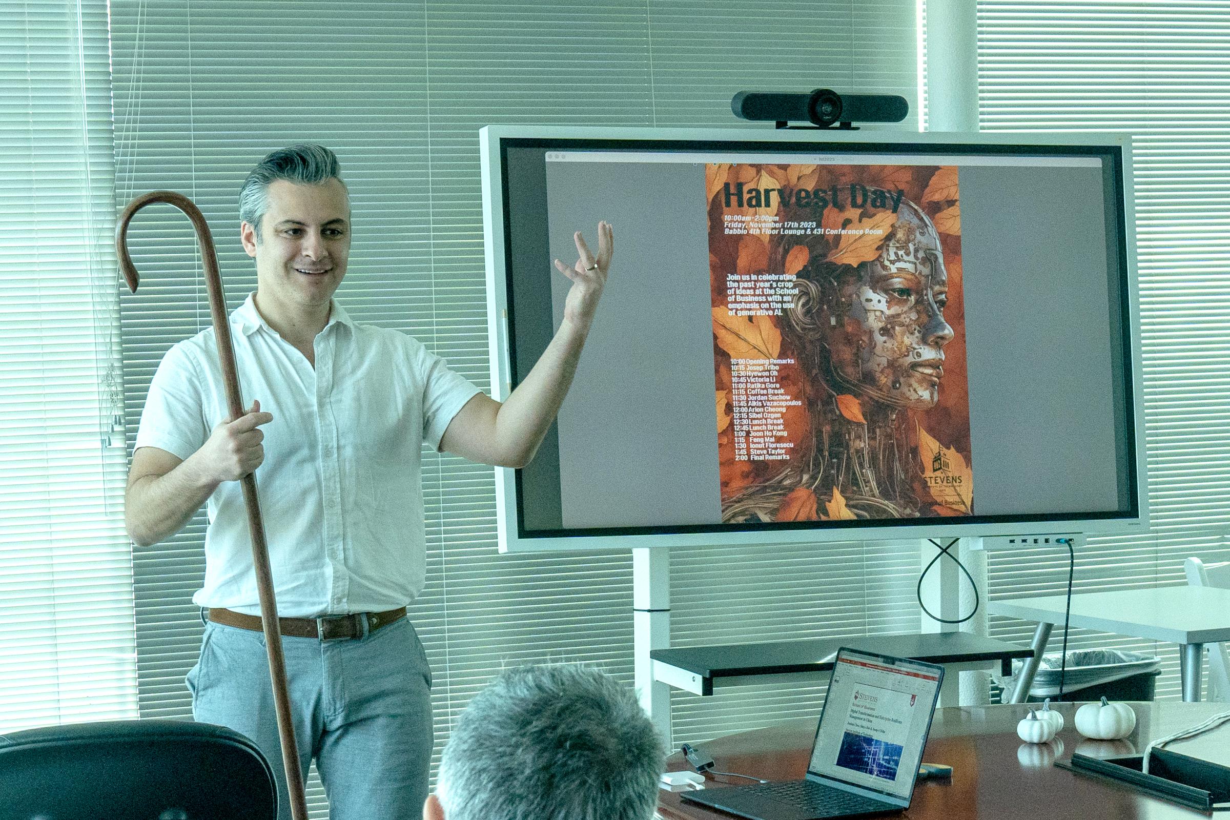 Jordan Suchow holds a shepherd's crook next to a smart board with an AI generated photo of a robot on a background of autumn leaves with the Harvest Day schedule on top.