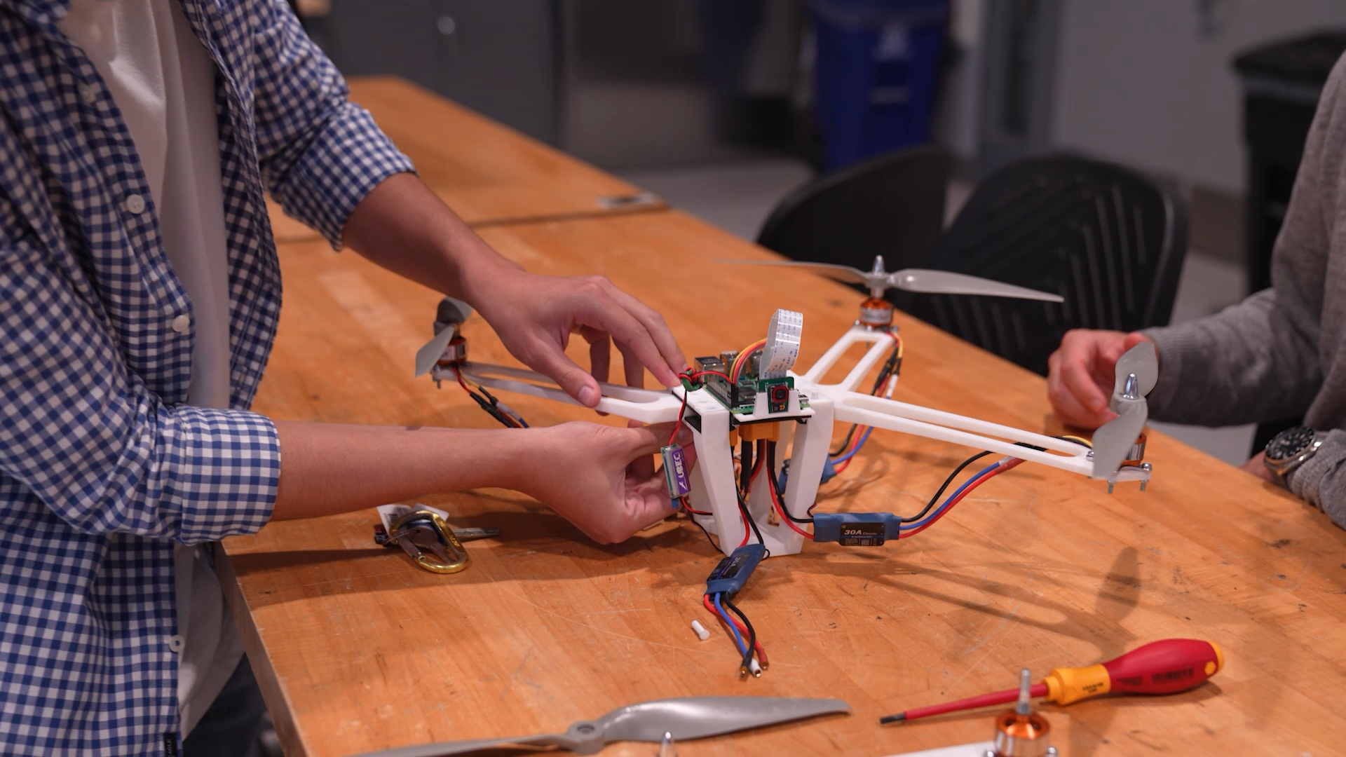 Two people collaborate on building a drone with white frame and exposed circuitry on a wooden workbench. Tools including pliers and screwdrivers are visible on the table. One person wears a blue checkered shirt while assembling the device.