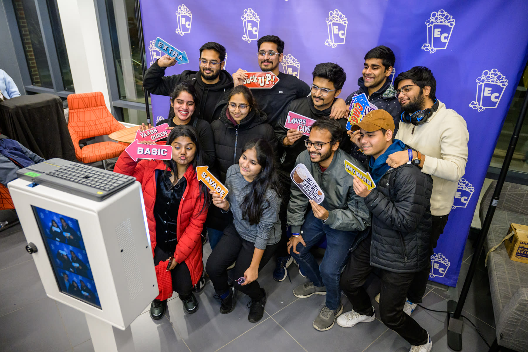 A group of students posing in front of a photo booth, holding up fun props featuring popular phrases like "Extra," "Basic," and "Savage." The props add a playful touch to the scene, with the students smiling and having fun.
