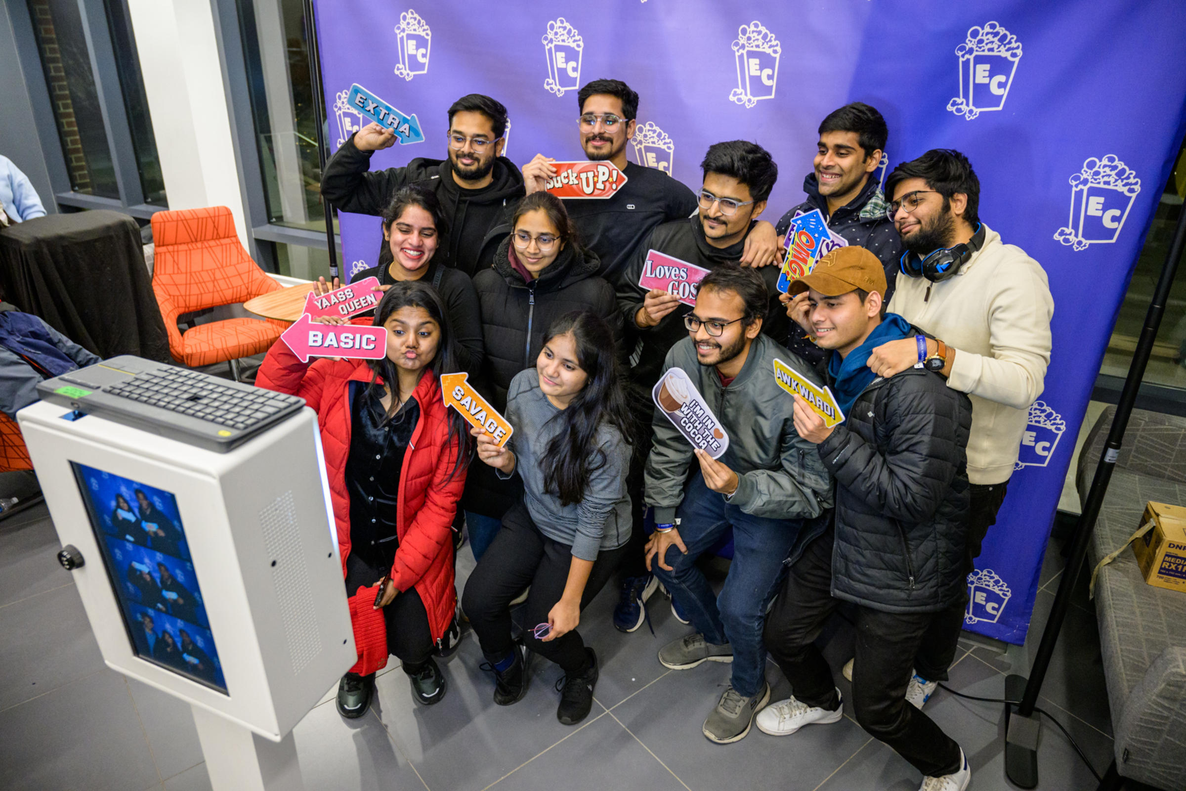 A group of students posing in front of a photo booth, holding up fun props featuring popular phrases like "Extra," "Basic," and "Savage." The props add a playful touch to the scene, with the students smiling and having fun.
