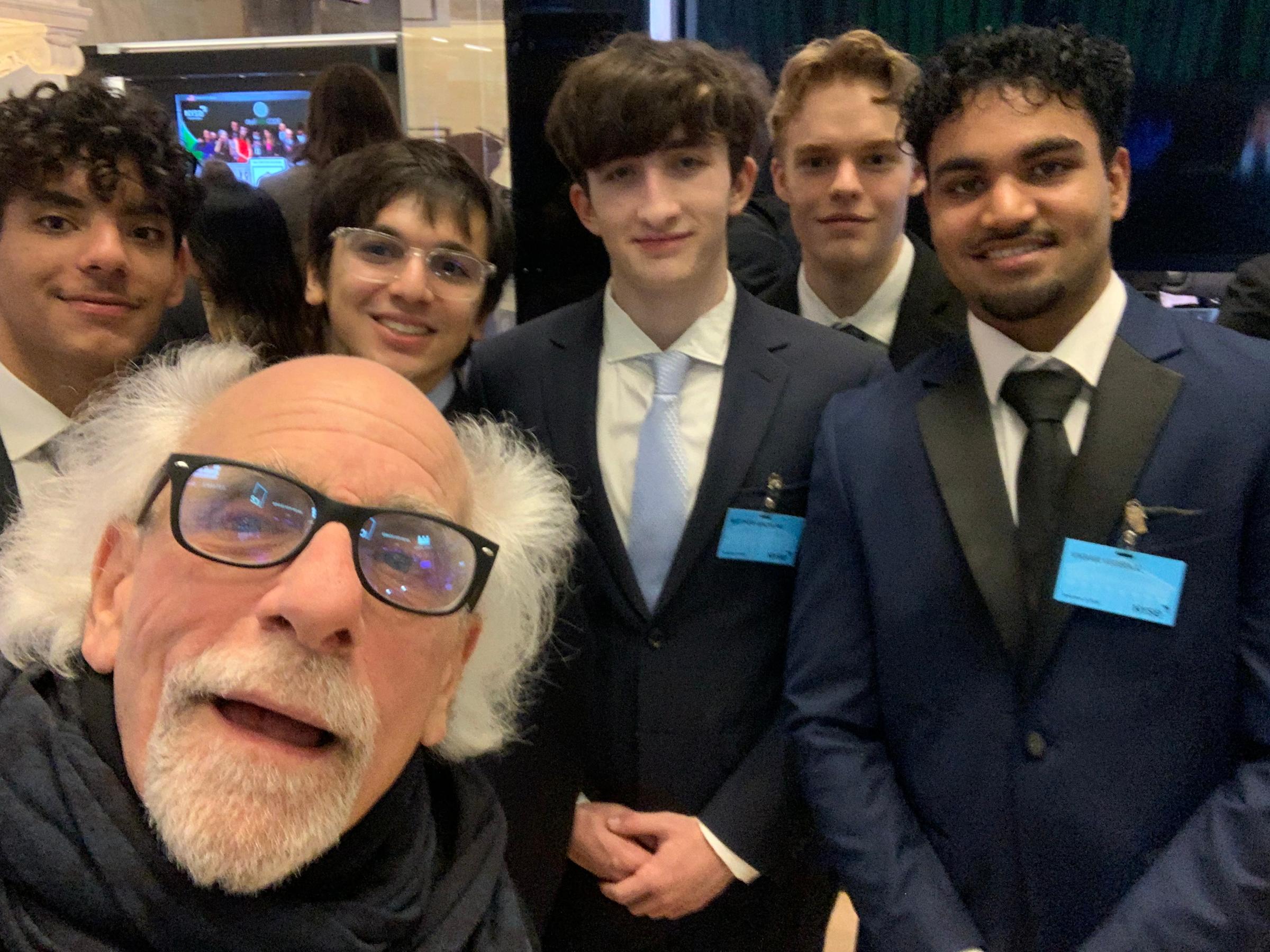 Selfie of Peter Tuchman with glasses and white hair and beard with five young male students in dark suits and ties wearing blue name badges at what appears to be a business event or competition.