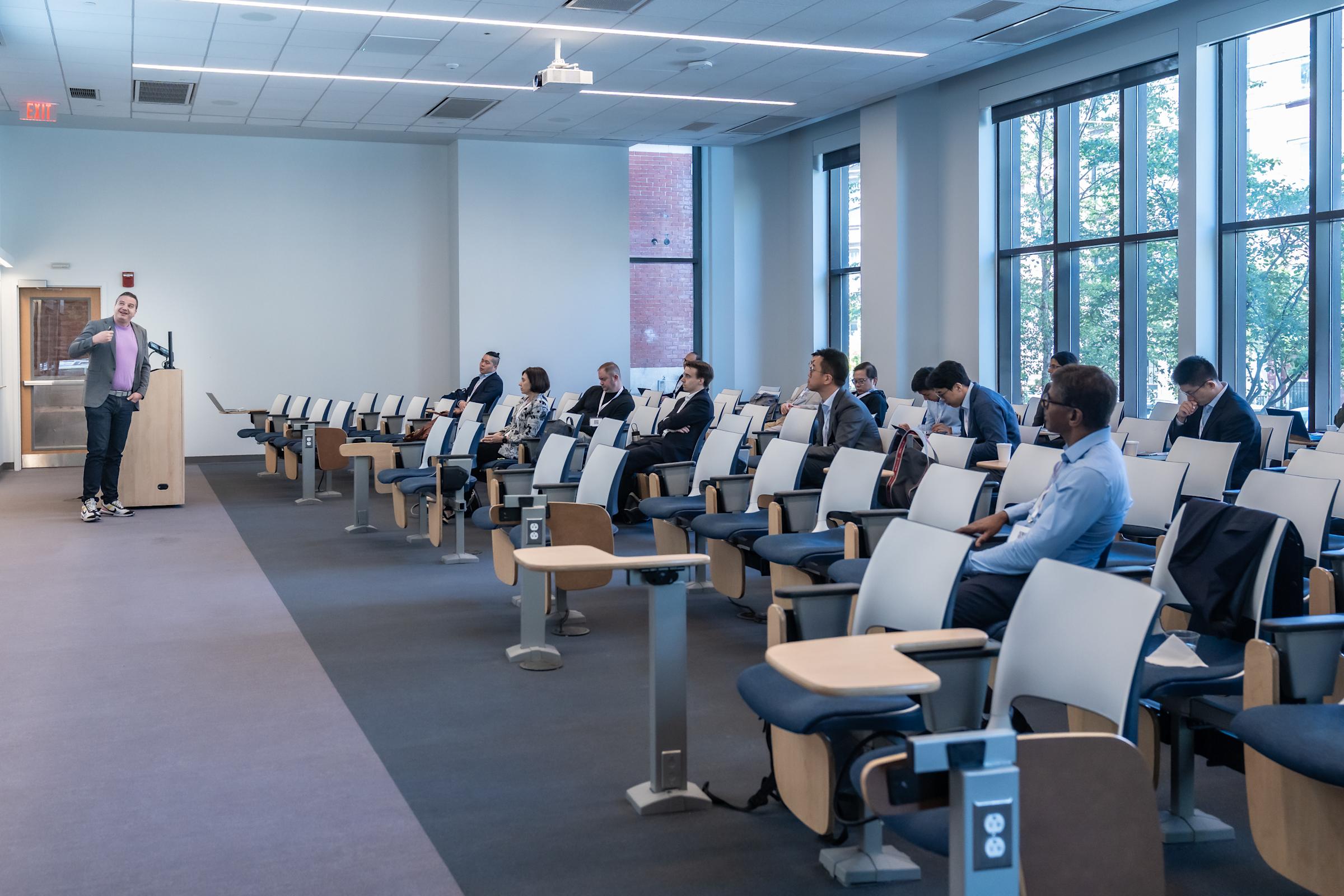 A presenter in casual attire speaks from a podium to an audience seated in modern tiered lecture hall seating. The bright room features large windows, white walls, and attendees in business attire listening to the presentation.