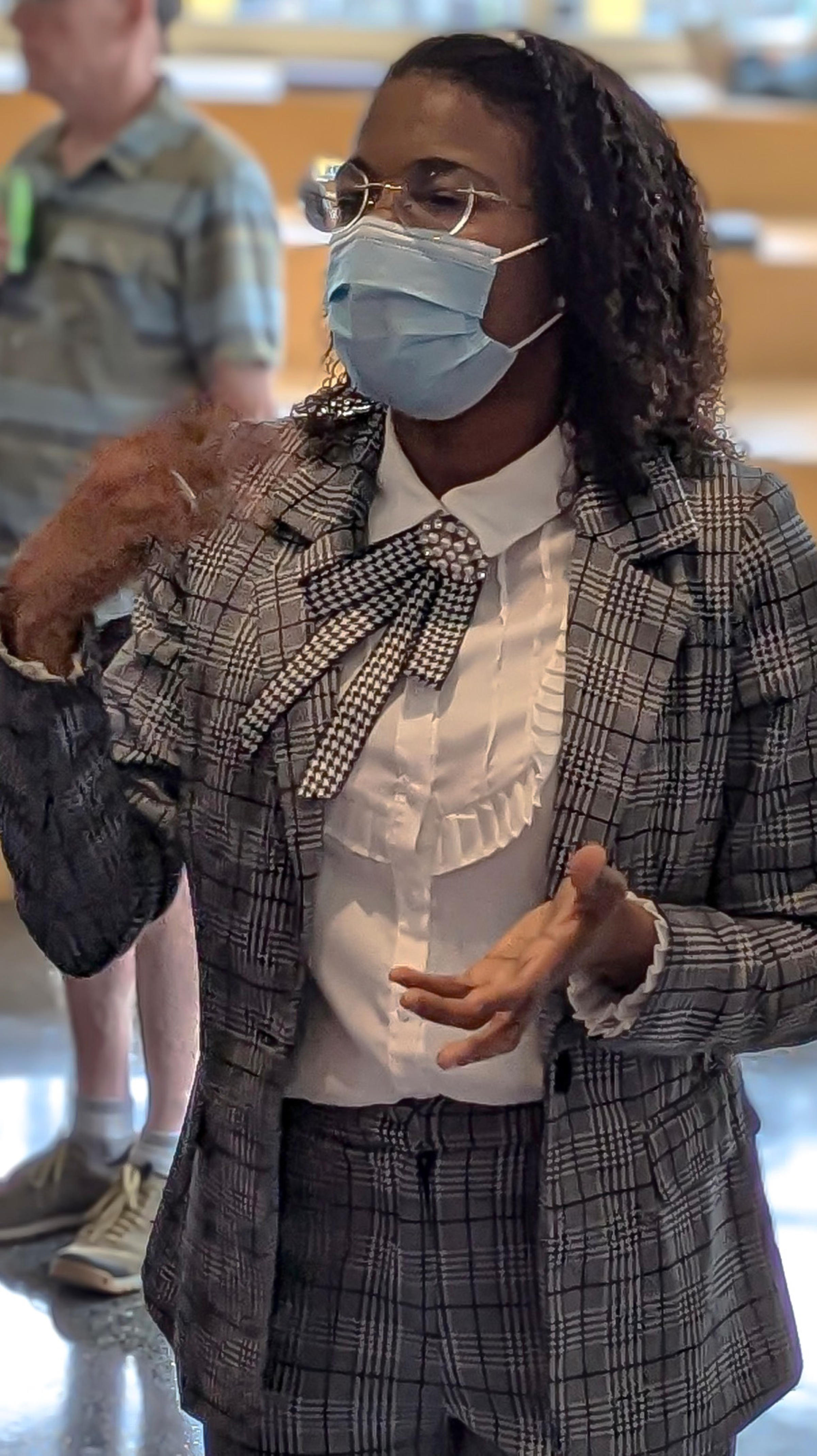 A woman with a surgeon's mask wearing a gray business suit speaking and gesturing with her hands.