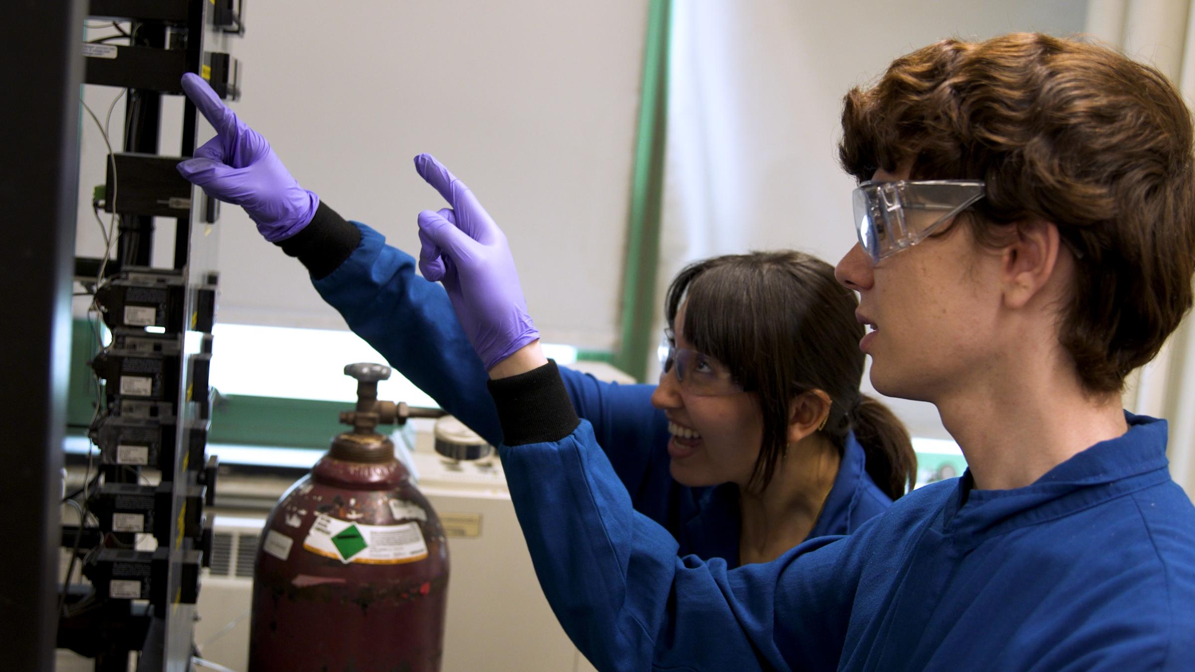 Two students in lab coats and latex gloves, pointing at something in the lab and smiling.