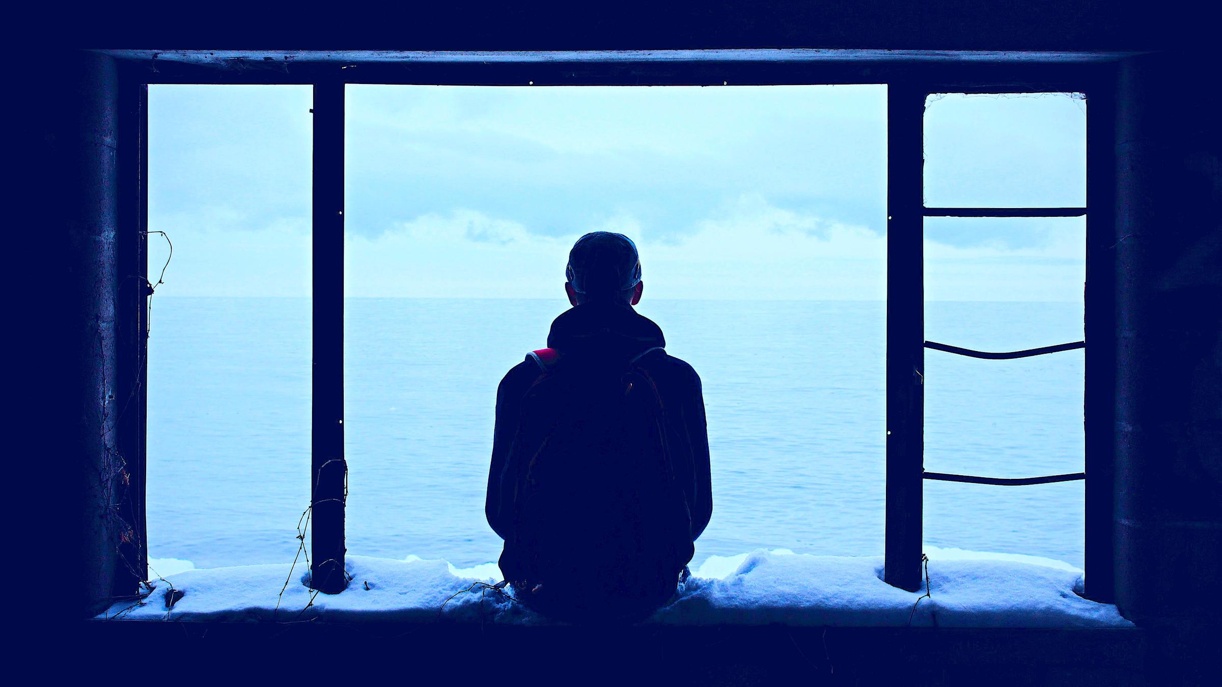 Silhouette of man staring out open window onto ocean, signifying loneliness