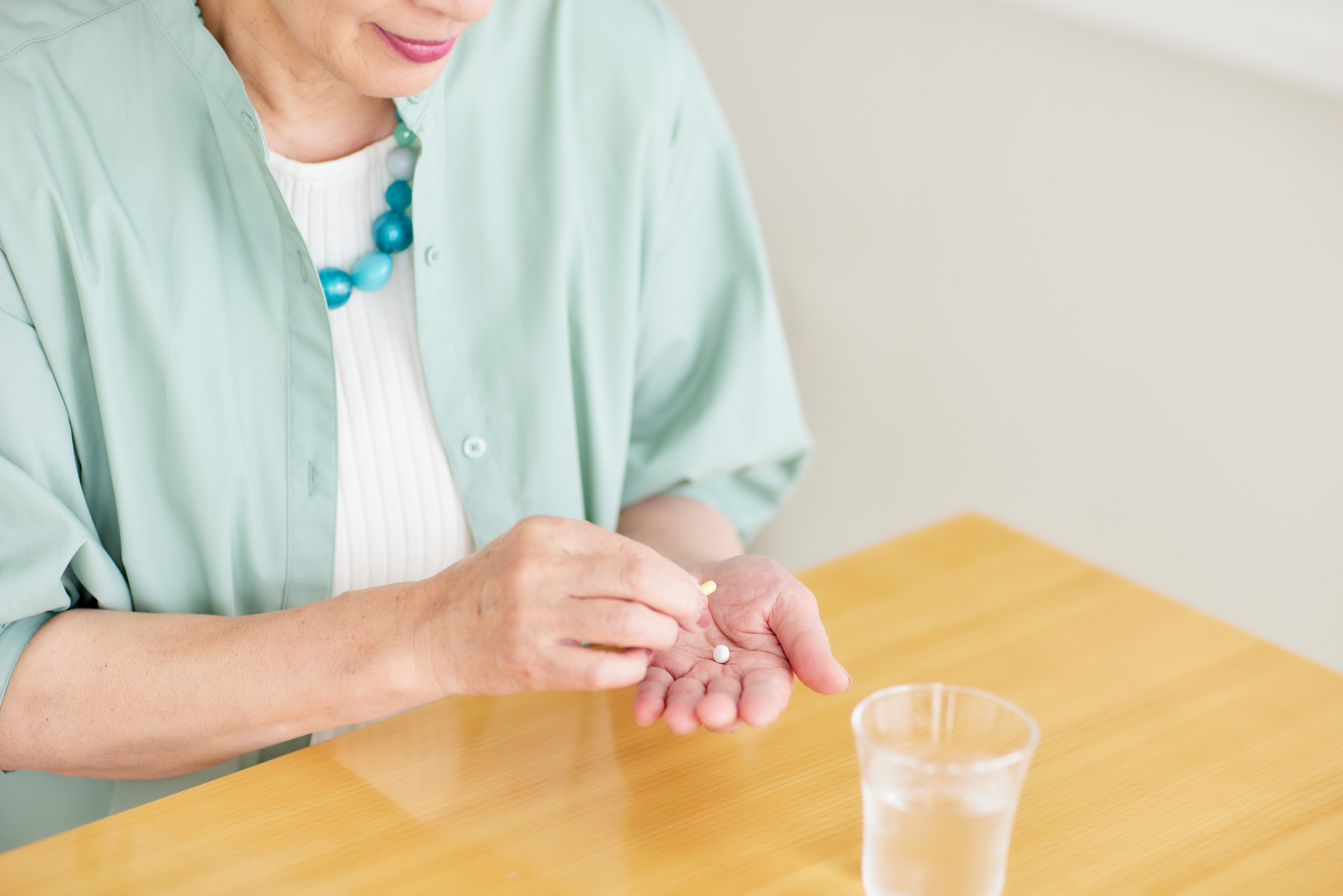 An elderly woman sits at a table holding a pill. There is a glass of water on the table.
