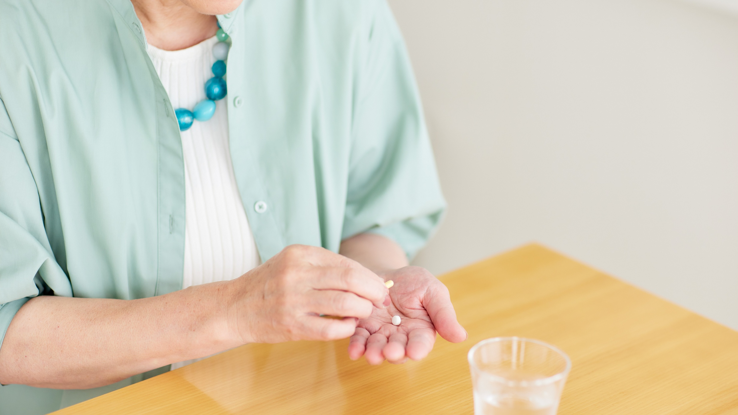 An elderly woman sits at a table holding a pill. There is a glass of water on the table.