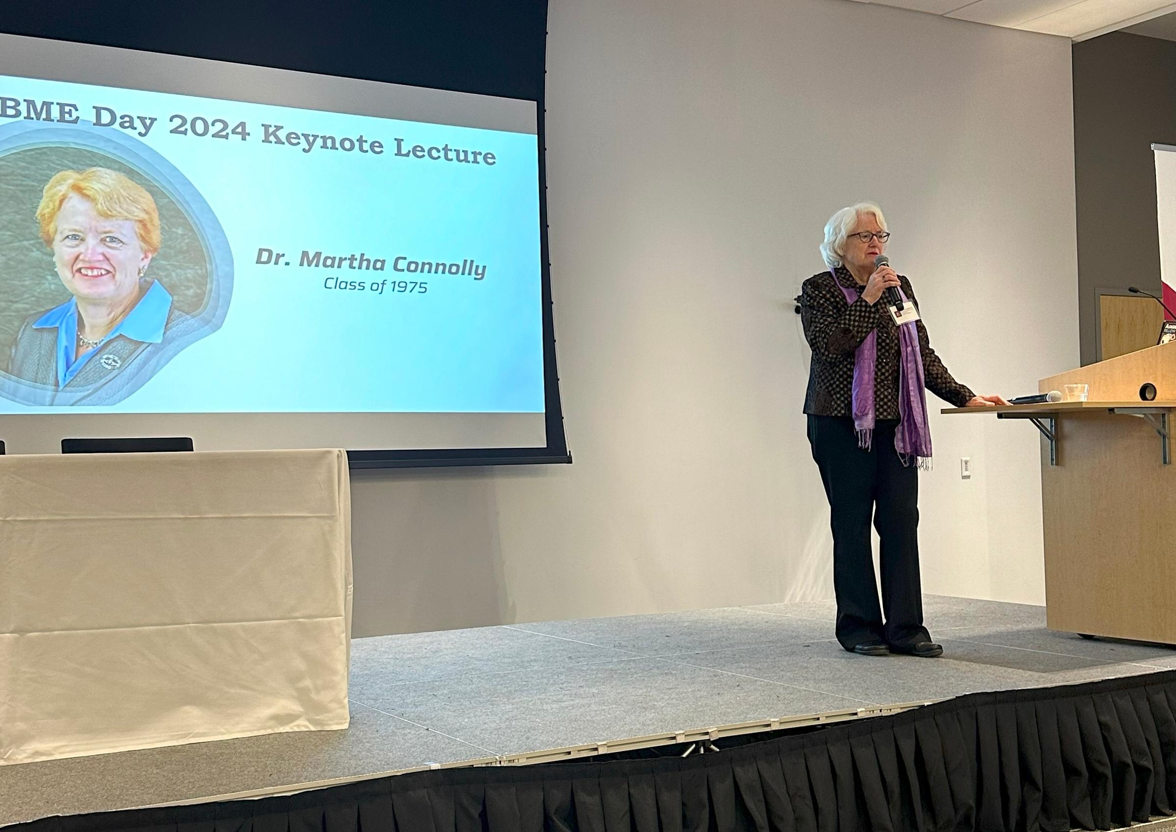 Martha Connolly stands at a podium in front a projector introducing the keynote lecture