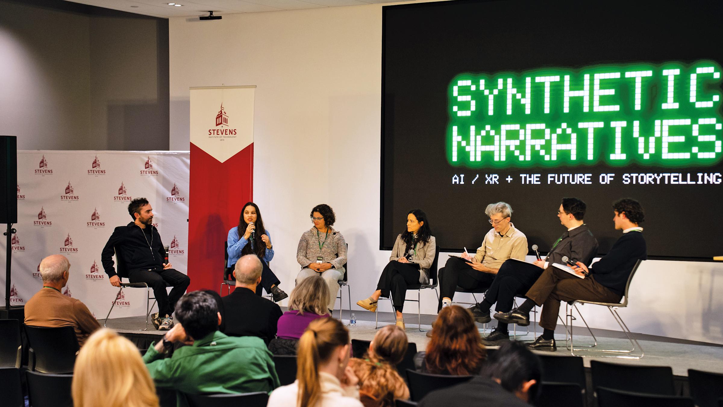 Photo of seven people gathered on a stage, one speaking, and facing an audience, with a fluorescent sign flashing “Synthetic Narratives” in the background.