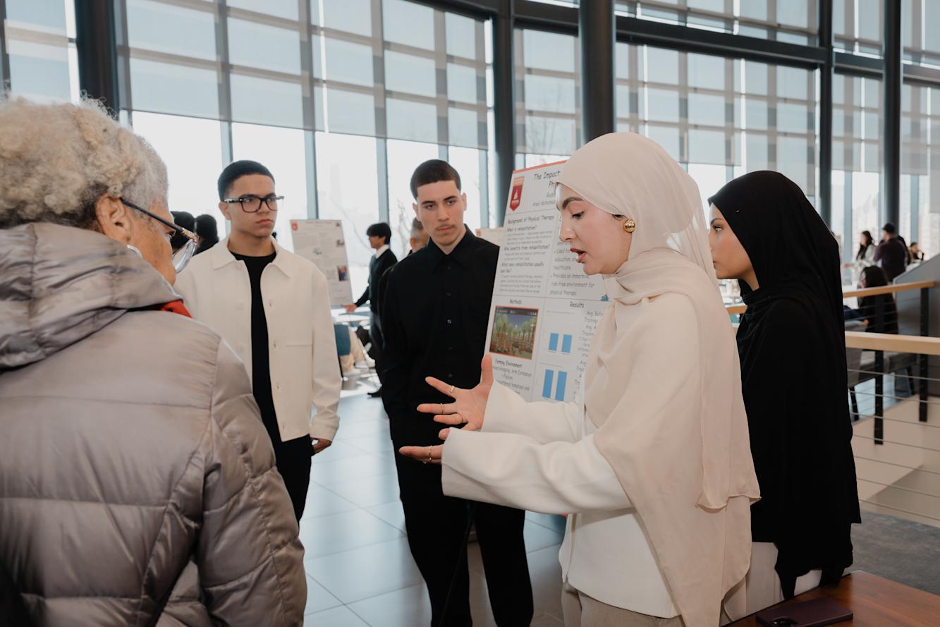 A female student in the foreground uses her hands to explain the research on her poster while two young men and an older woman listen.