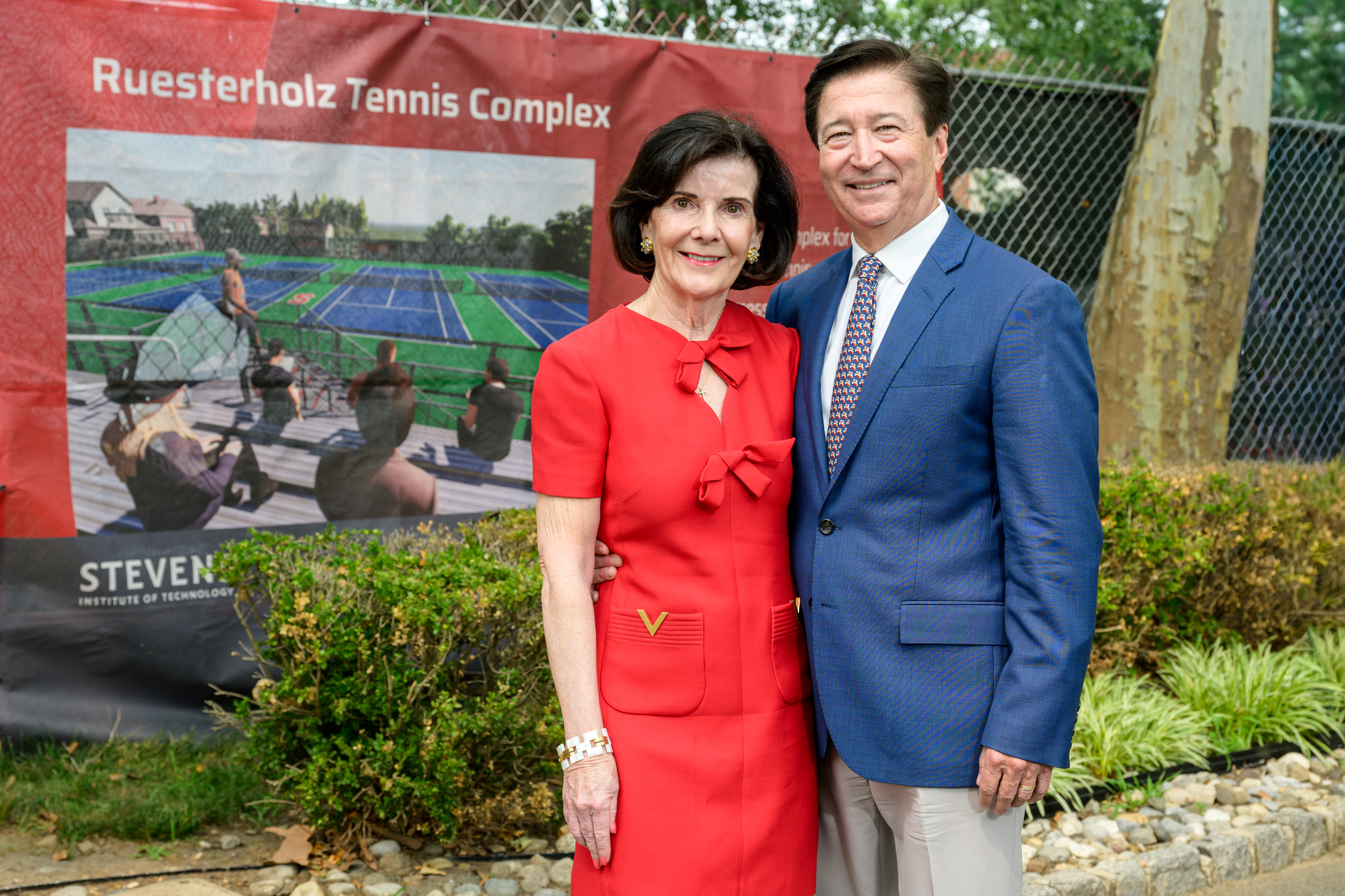 Virginia Ruesterholz ’83 Hon. D.Eng. ’08 and Kevin Ruesterholz ’83 stand in front of a Ruesterholz Tennis Complex sign displaying a rendering of new tennis courts at Stevens Institute of Technology. Virginia wears a red dress and Kevin wears a blue blazer.