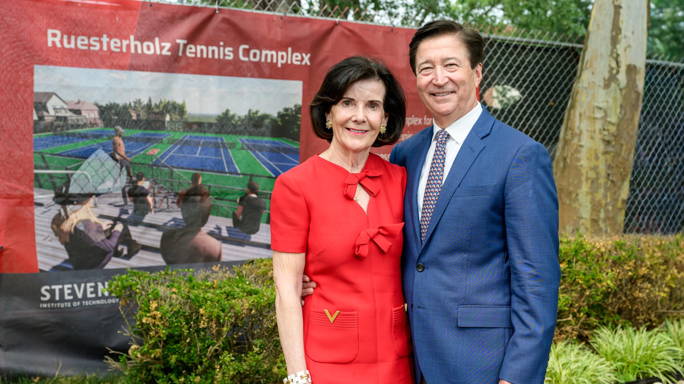 Virginia Ruesterholz ’83 Hon. D.Eng. ’08 and Kevin Ruesterholz ’83 stand in front of a Ruesterholz Tennis Complex sign displaying a rendering of new tennis courts at Stevens Institute of Technology. Virginia wears a red dress and Kevin wears a blue blazer.
