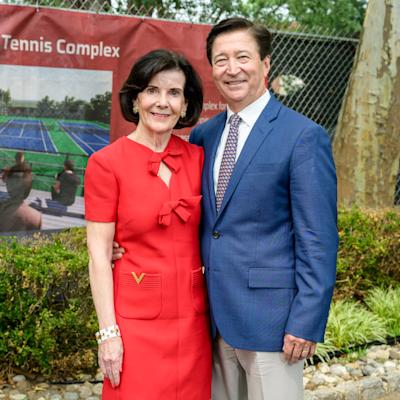 Virginia Ruesterholz ’83 Hon. D.Eng. ’08 and Kevin Ruesterholz ’83 stand in front of a Ruesterholz Tennis Complex sign displaying a rendering of new tennis courts at Stevens Institute of Technology. Virginia wears a red dress and Kevin wears a blue blazer.