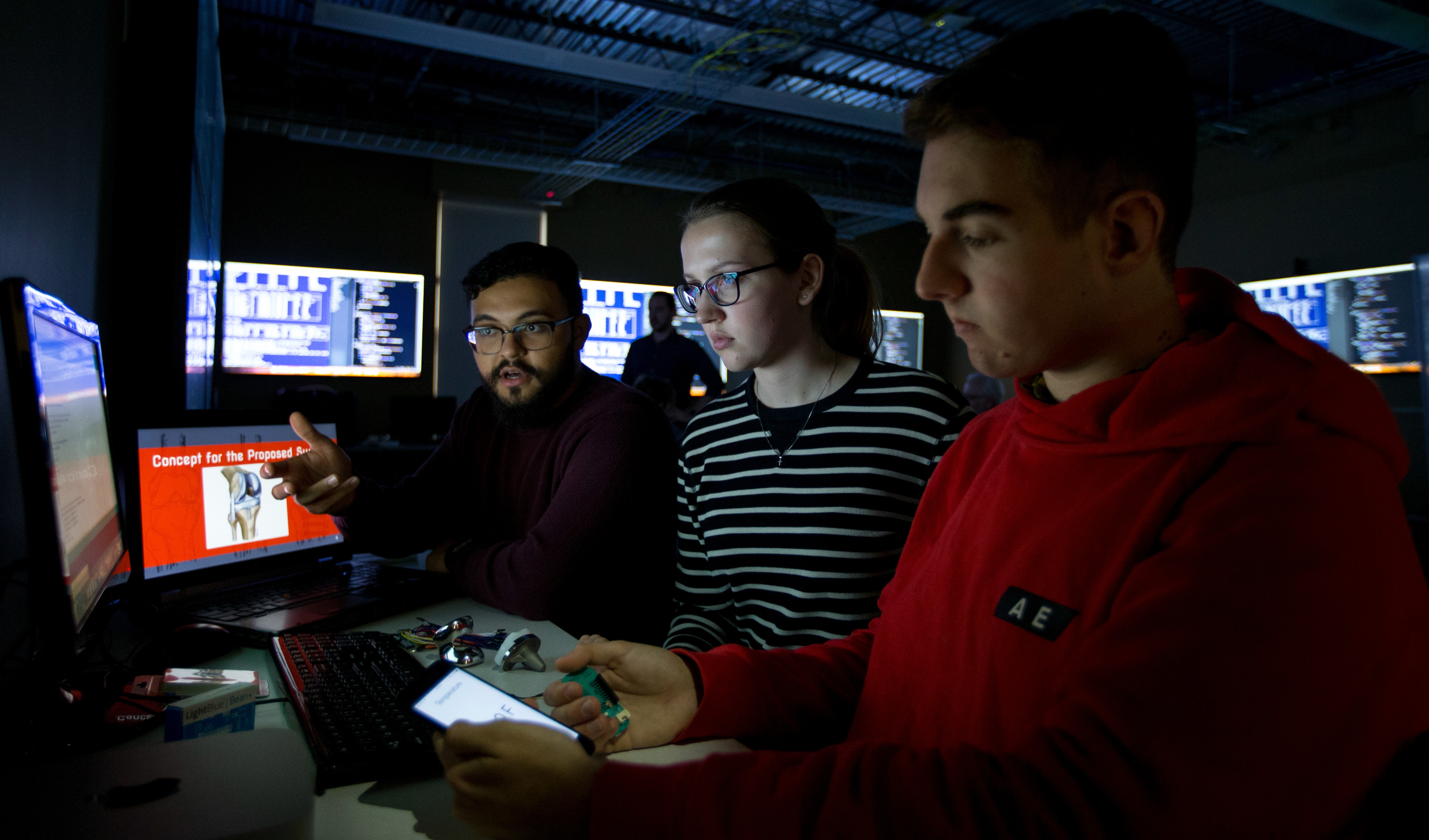 Three students sitting in a dark room surrounded by monitors. 