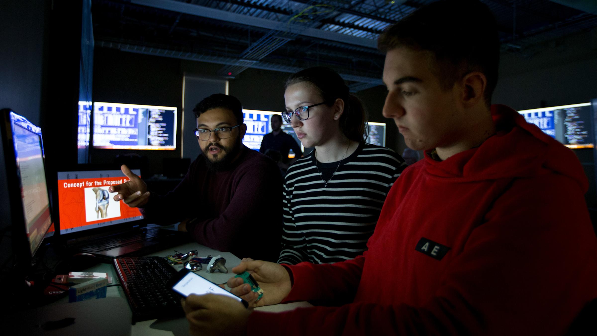Three students sitting in a dark room surrounded by monitors.