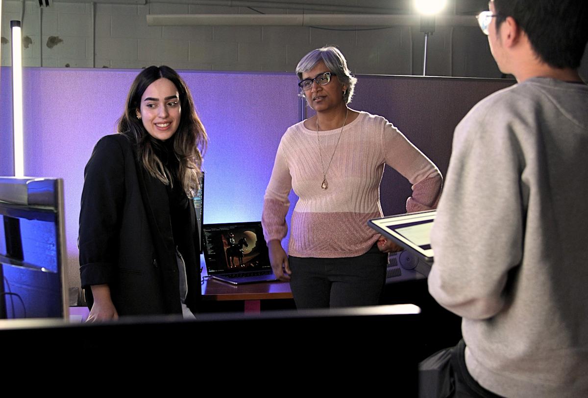 A photograph of K.P. “Suba” Subbalakshmi standing in the middle of two research students, looking at a computer screen under blue lighting.