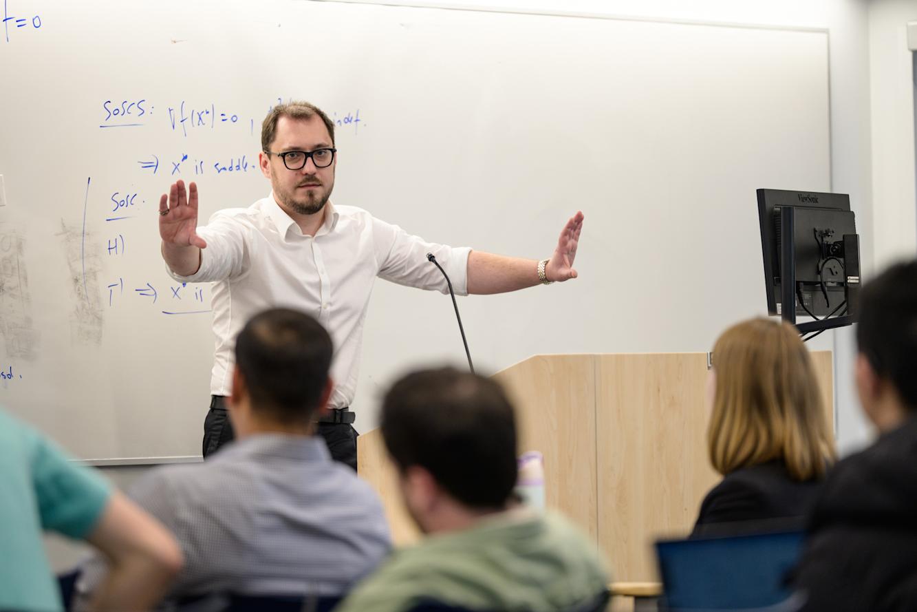 A man standing at a speaking podium giving a talk and gesturing to a group of mathematical sciences students.