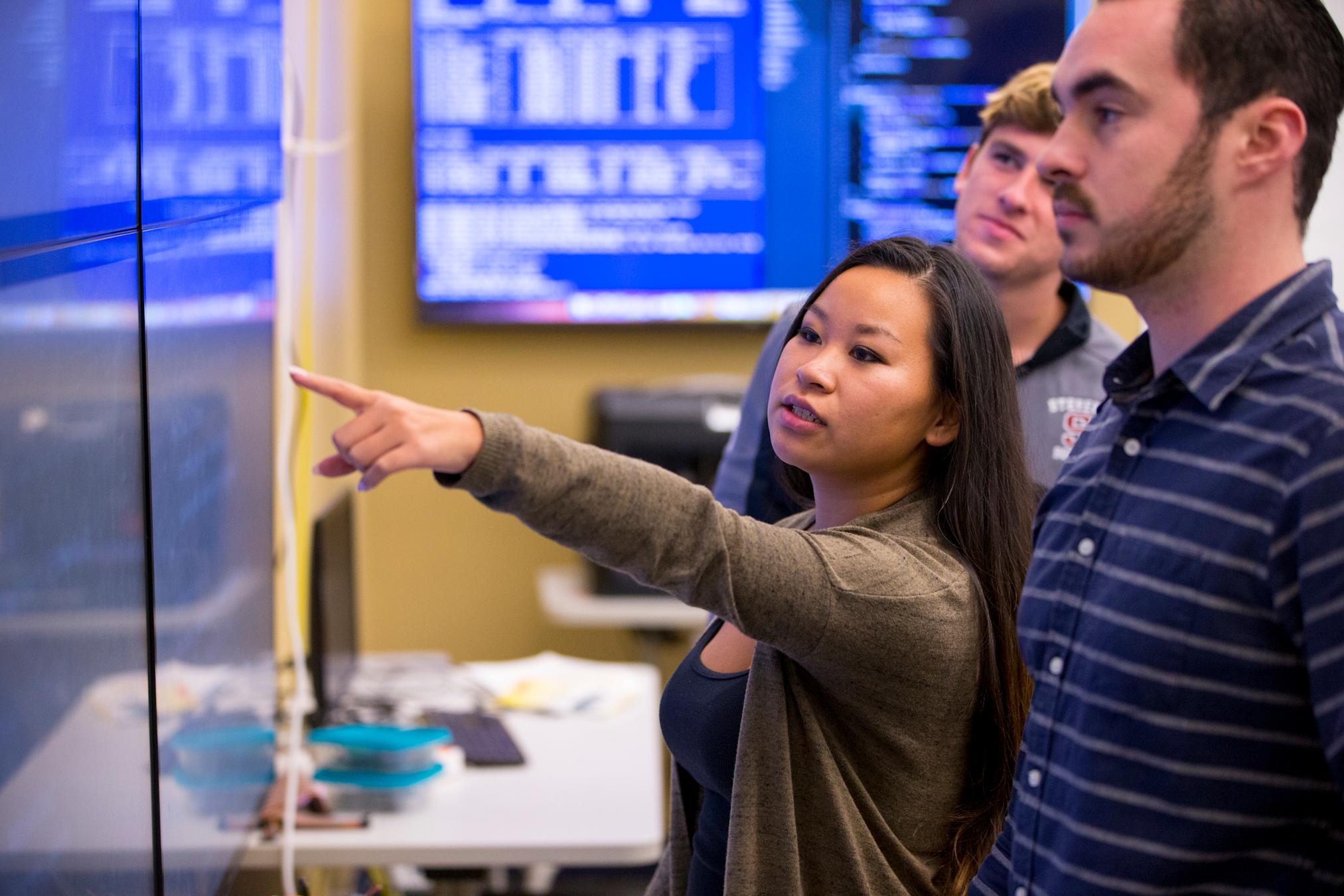 Students standing in class analyzing code on wall screen