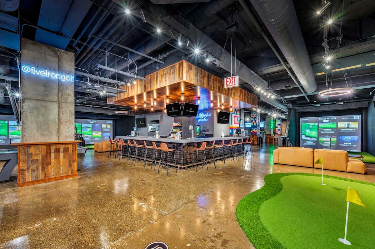 Interior view of Five Iron Golf in New York City, featuring a central bar, indoor putting green, golf simulators, lounge seating and modern industrial décor.