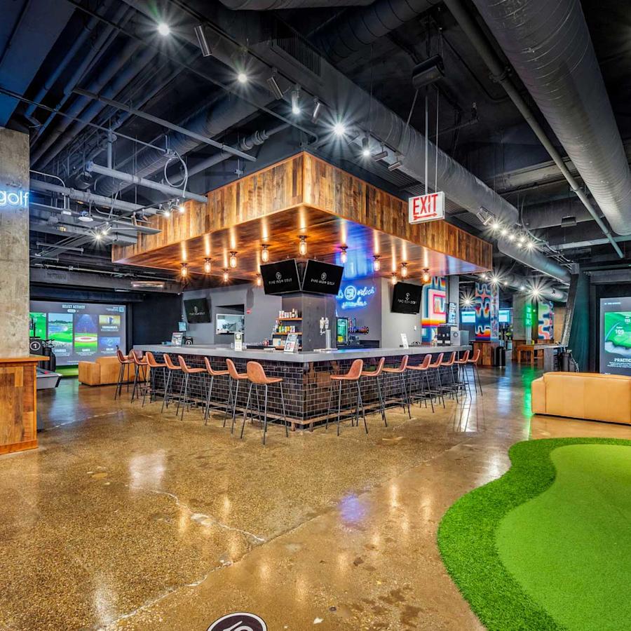 Interior view of Five Iron Golf in New York City, featuring a central bar, indoor putting green, golf simulators, lounge seating and modern industrial décor.