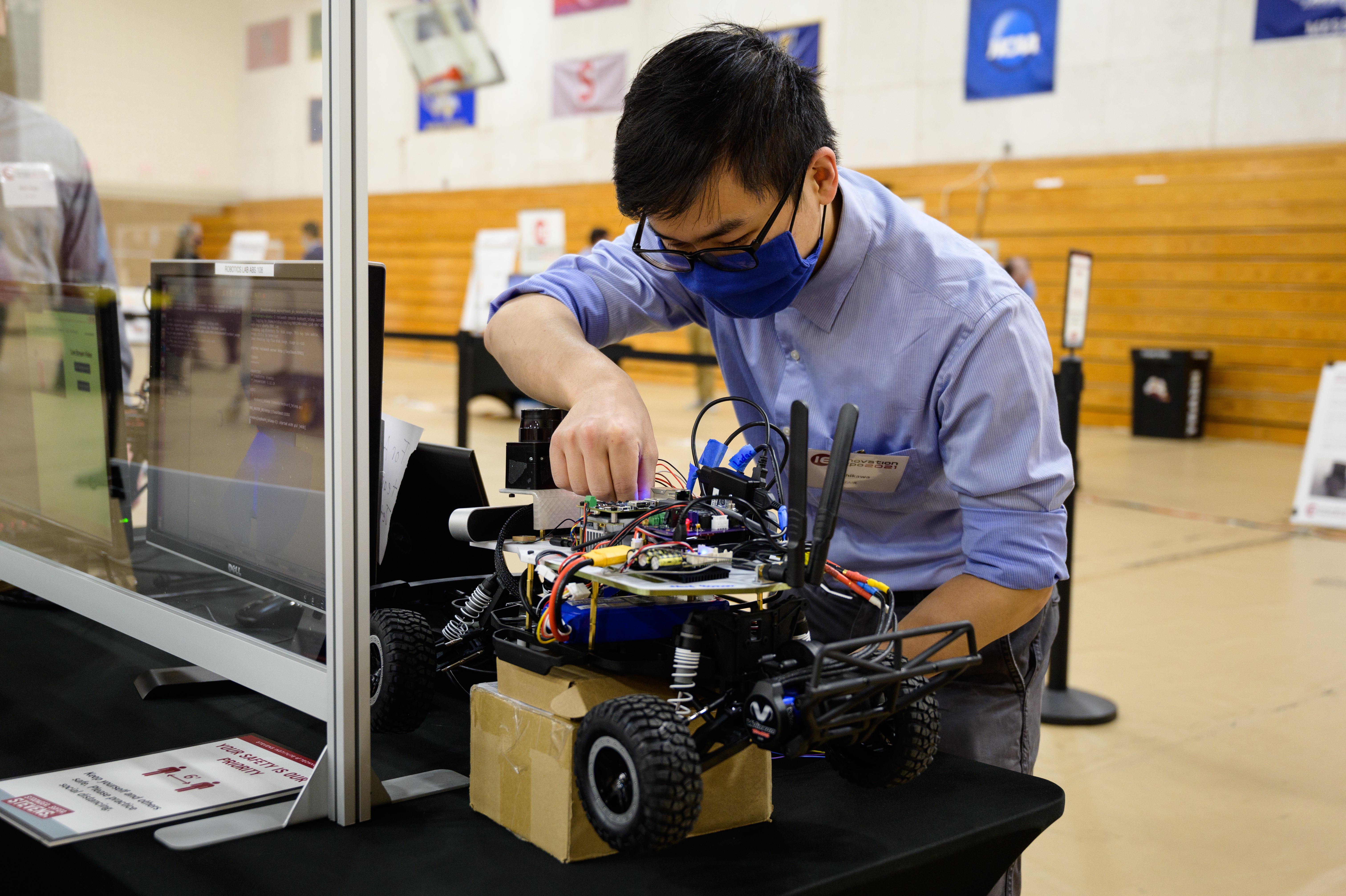 A student wearing a black mask and blue shirt. He is leaning over a robotic vehicle with wheels and adjusting some components on the top of it.