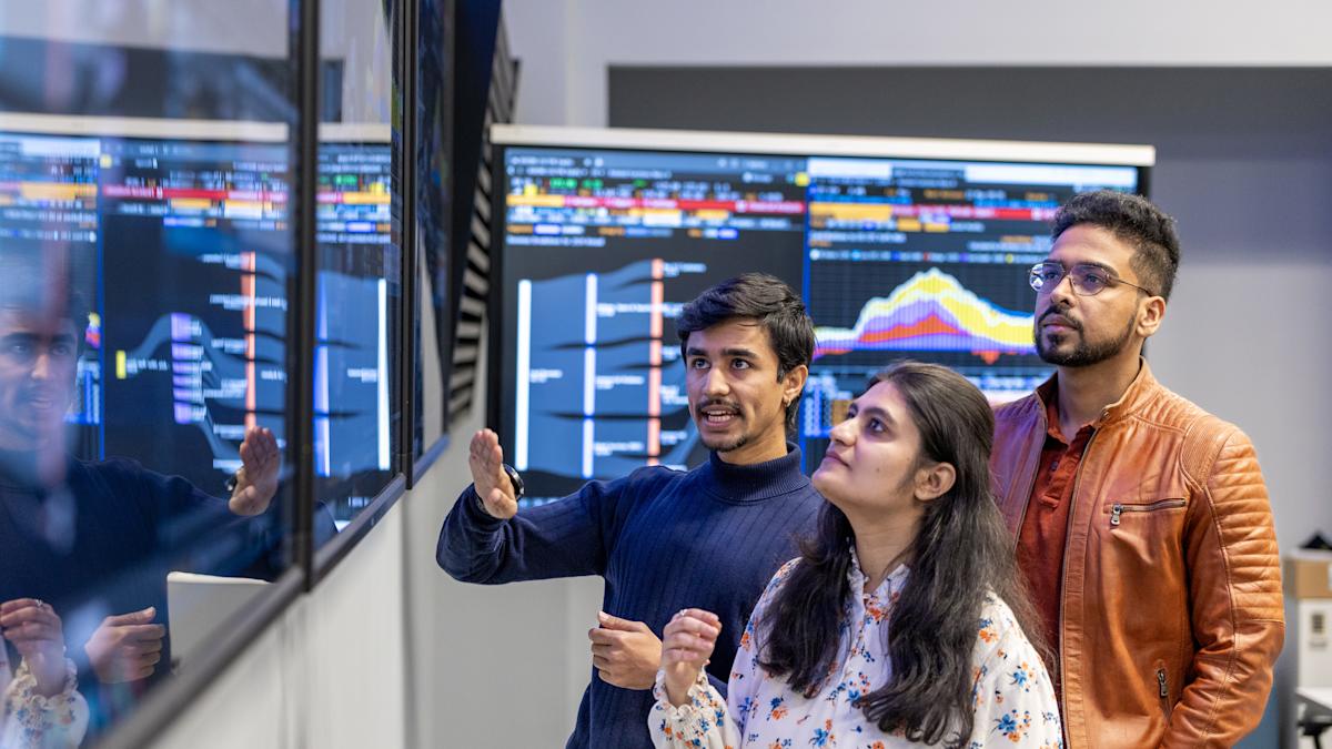 Three students discuss data displayed on a large screen in front of them.