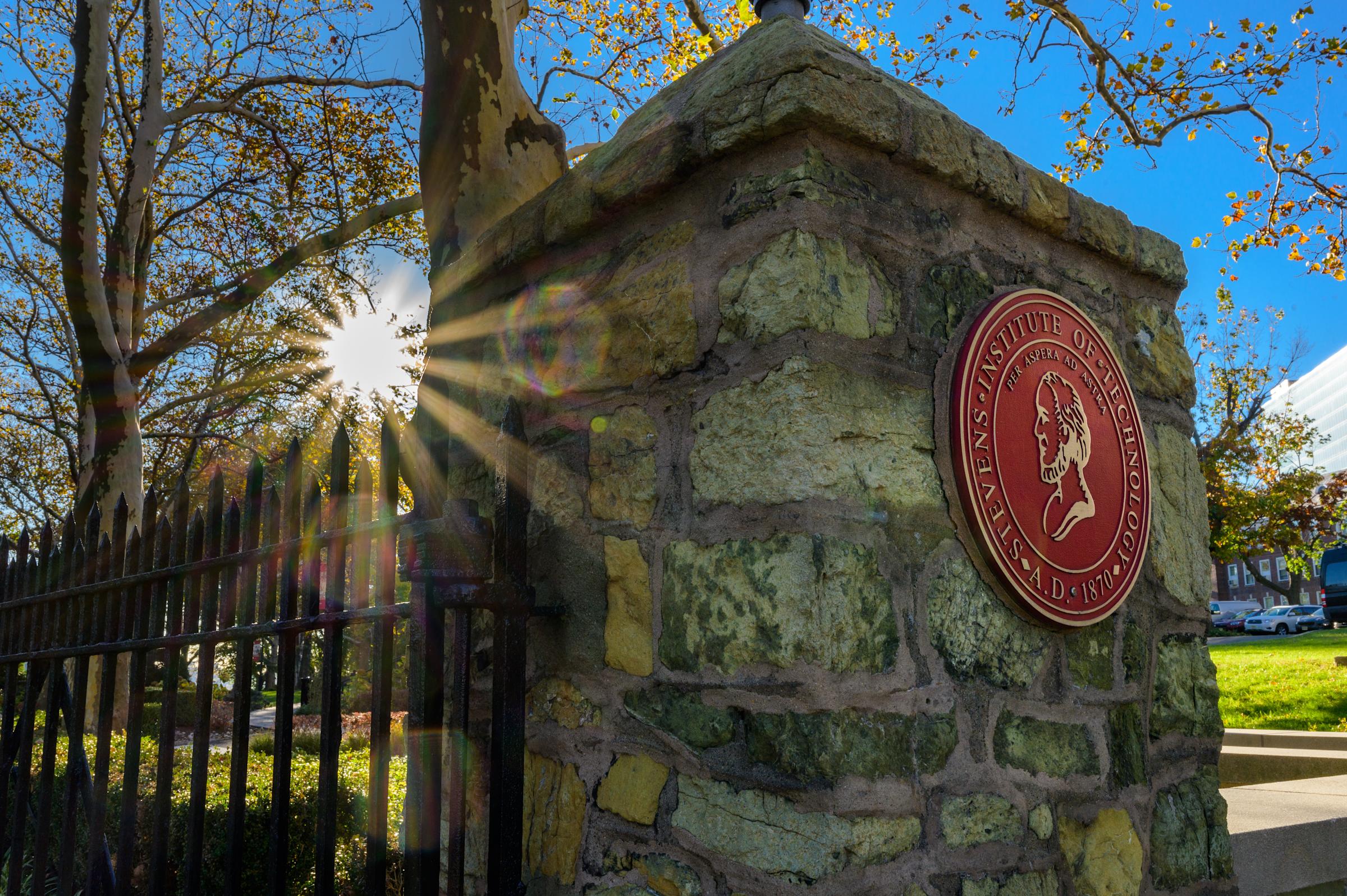 Gateway entrance to Stevens campus with presidents seal and sun shining through trees.