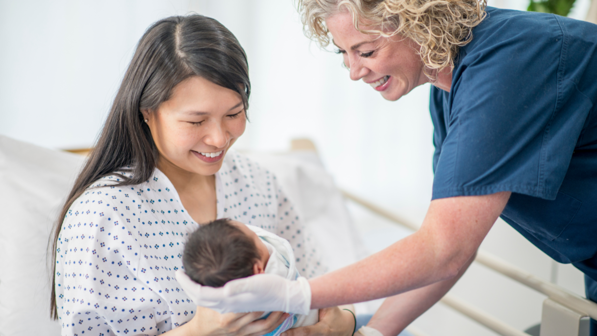 A mother sits up in a hospital bed holding a newborn baby. A female doctor leans over to help her from the foot of the bed.