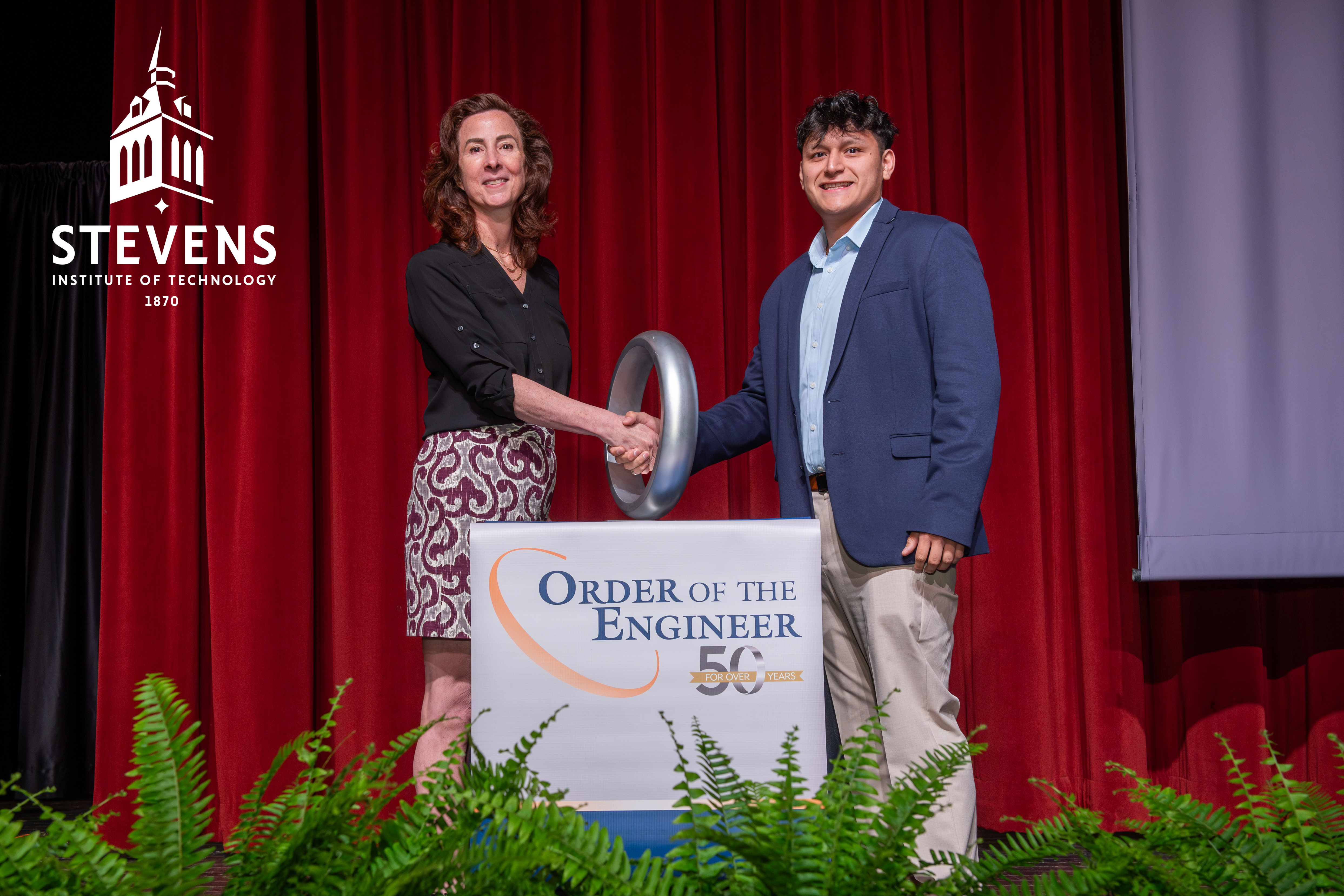 Two people stand behind a podium wearing business clothes. On the podium it reads "Order of the Engineer." The two people are shaking hands through a large ring that is on the podium.