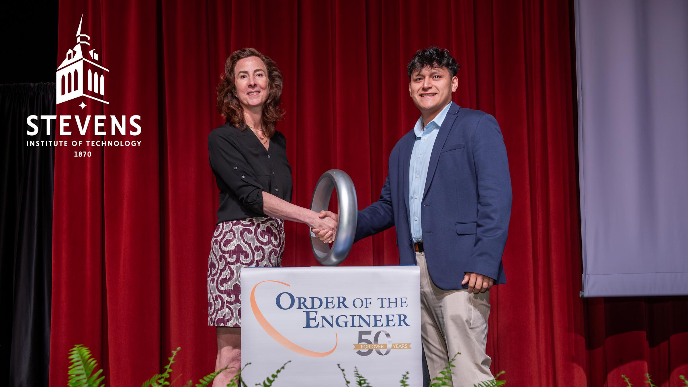 Two people stand behind a podium wearing business clothes. On the podium it reads "Order of the Engineer." The two people are shaking hands through a large ring that is on the podium.