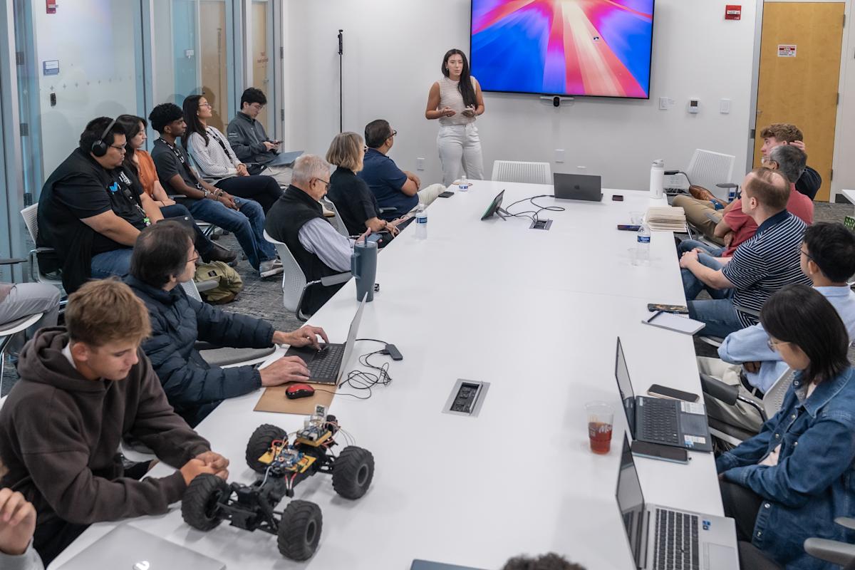 A woman presents beside a large wall screen as about 15 attendees listen around a long white conference table with laptops and water bottles. A homemade robotic RC car with exposed wiring sits on the table in the foreground.