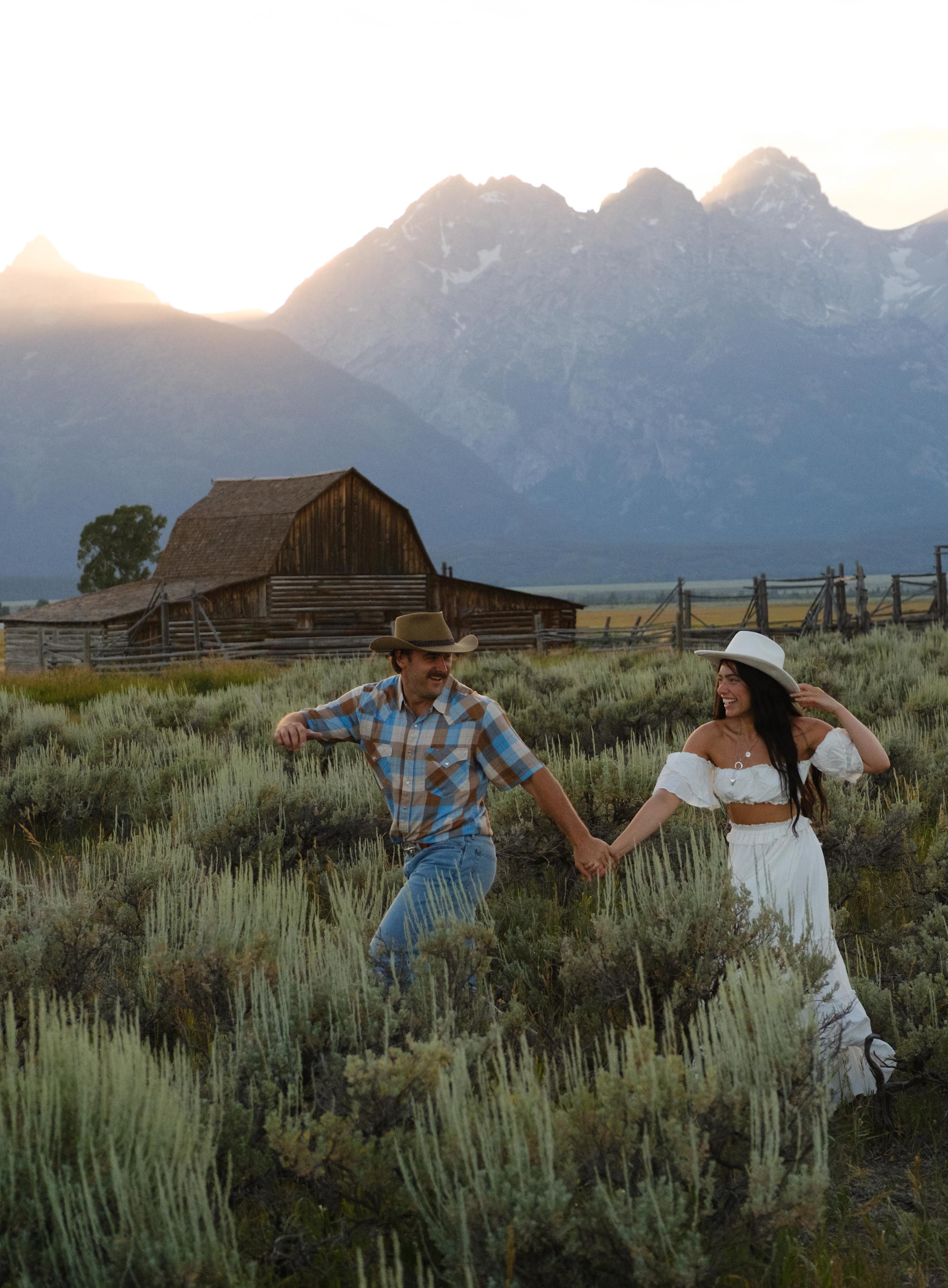 A couple walks hand-in-hand through sagebrush at sunset, both wearing cowboy hats. A historic wooden barn and the Grand Teton mountain range glow in warm golden light behind them, likely Mormon Row in Grand Teton National Park, Wyoming.