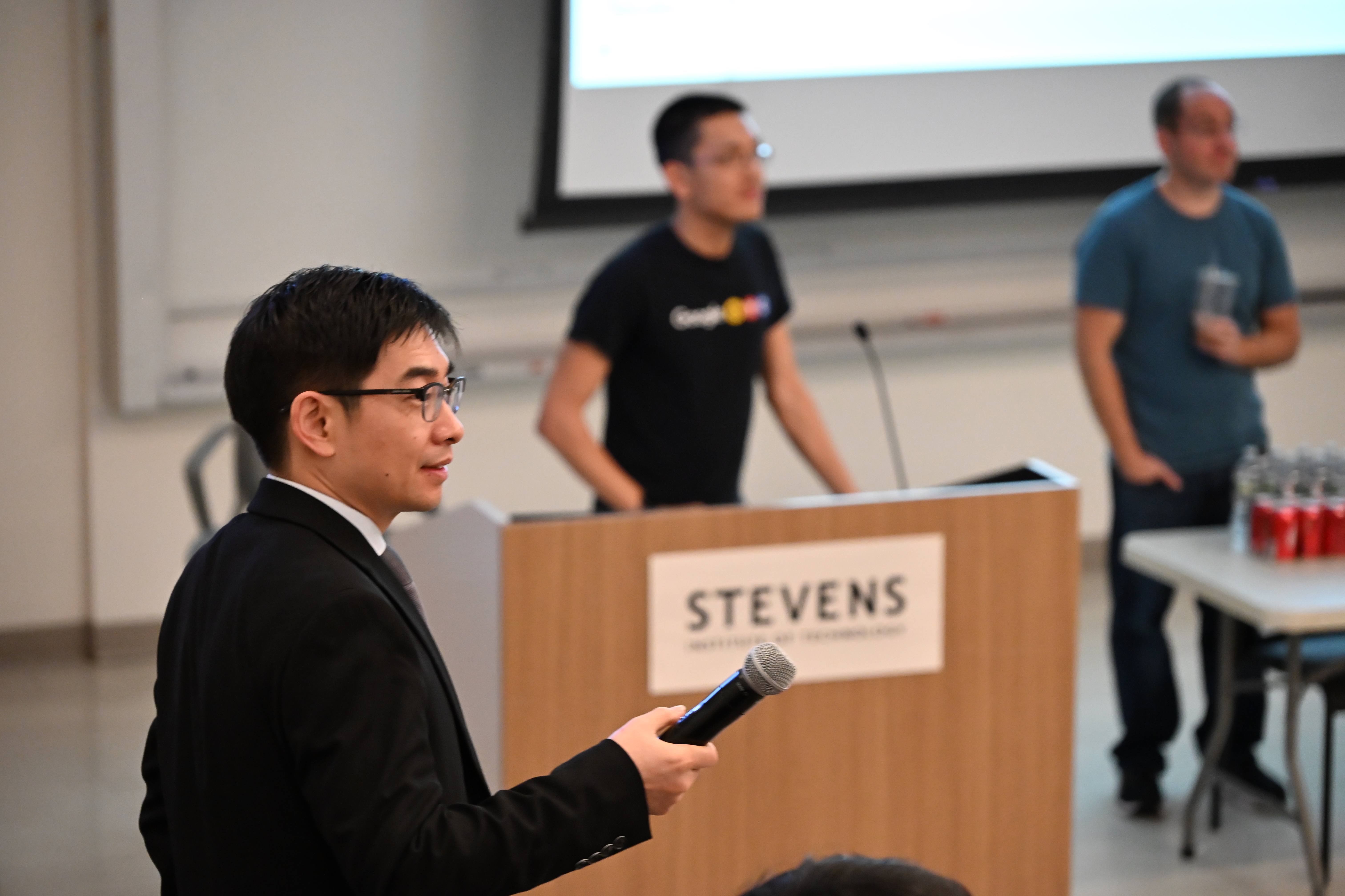 A photo of a man in a black suit holding a microphone on the left, with two figures of people behind a table and podium in the background.