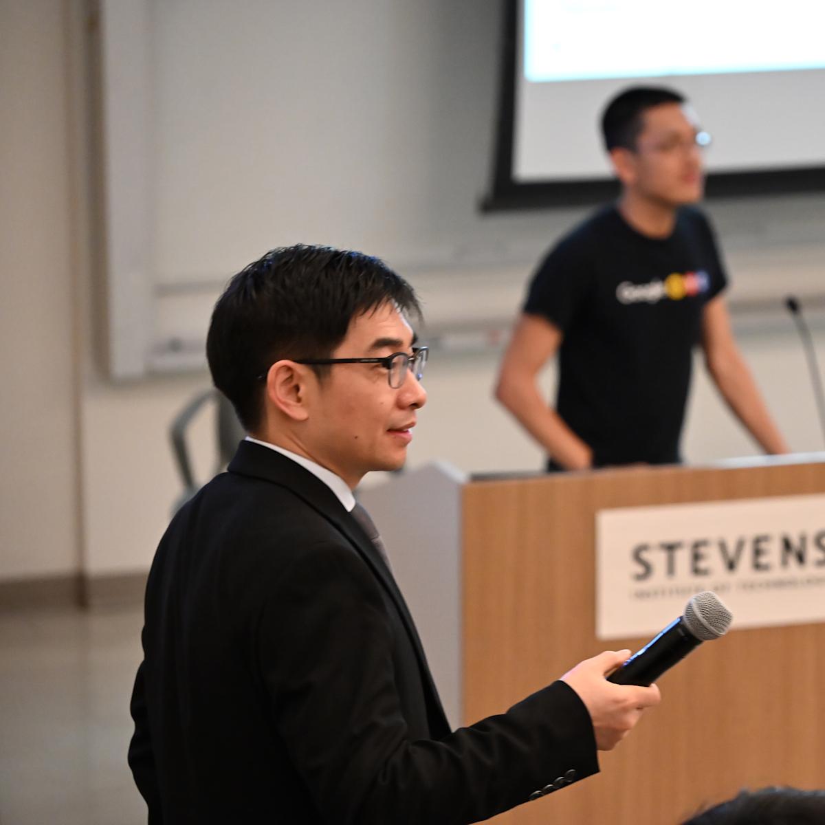 A photo of a man in a black suit holding a microphone on the left, with two figures of people behind a table and podium in the background.