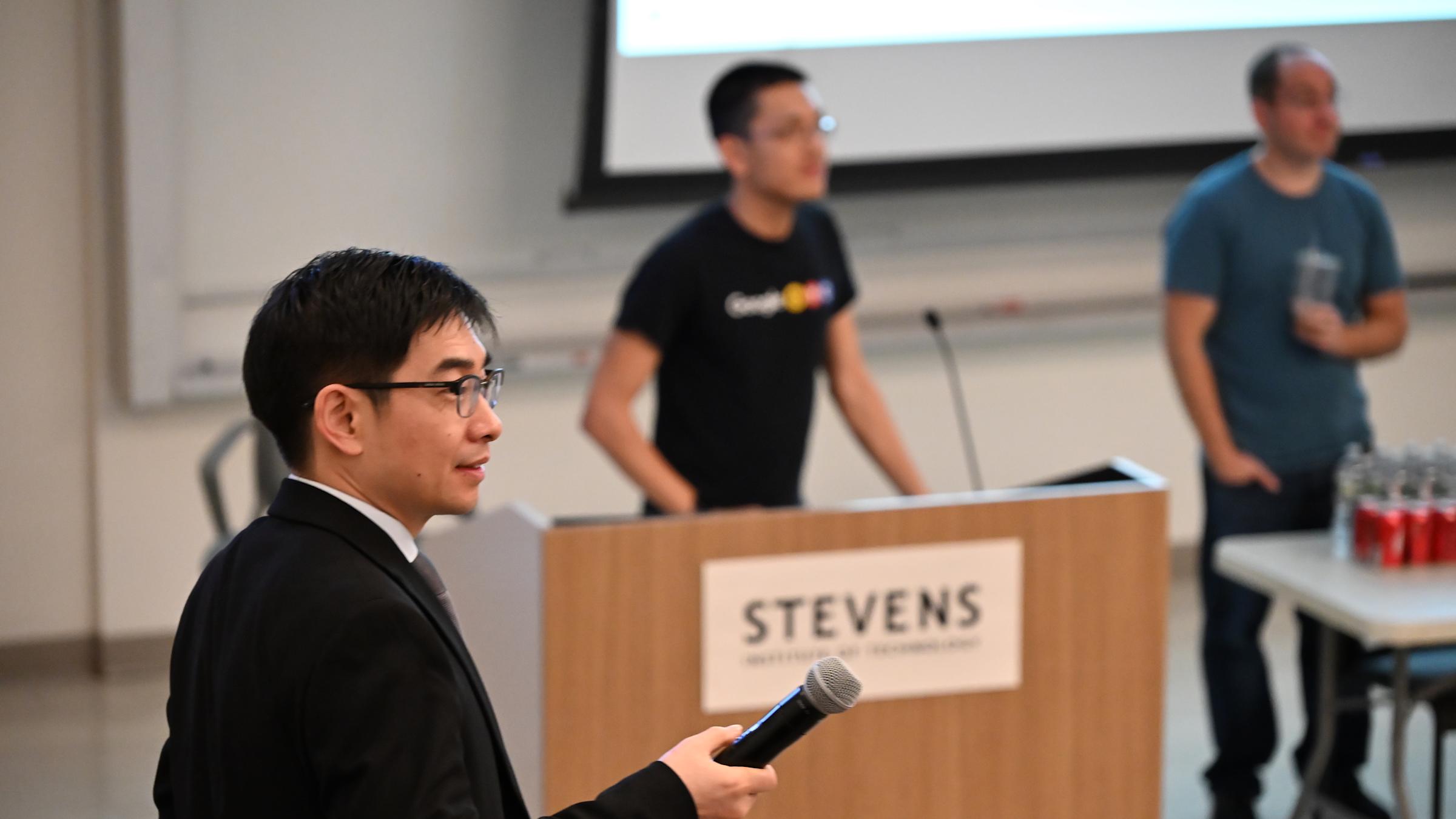 A photo of a man in a black suit holding a microphone on the left, with two figures of people behind a table and podium in the background.