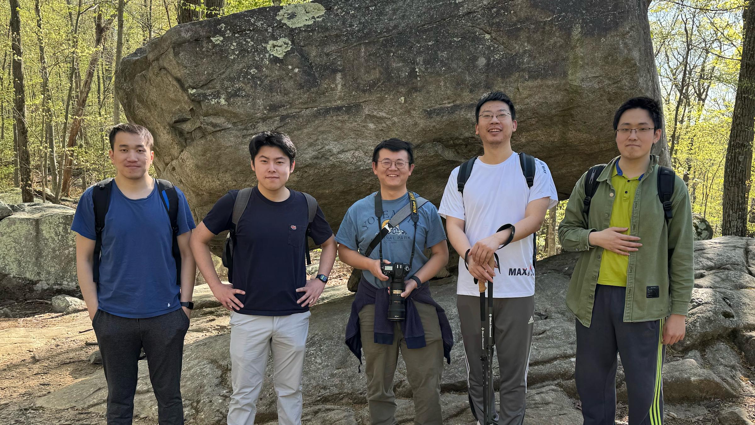 Hao Wang with two male Ph.D. students on each side of him pose on a hiking trail in front of a large rock.
