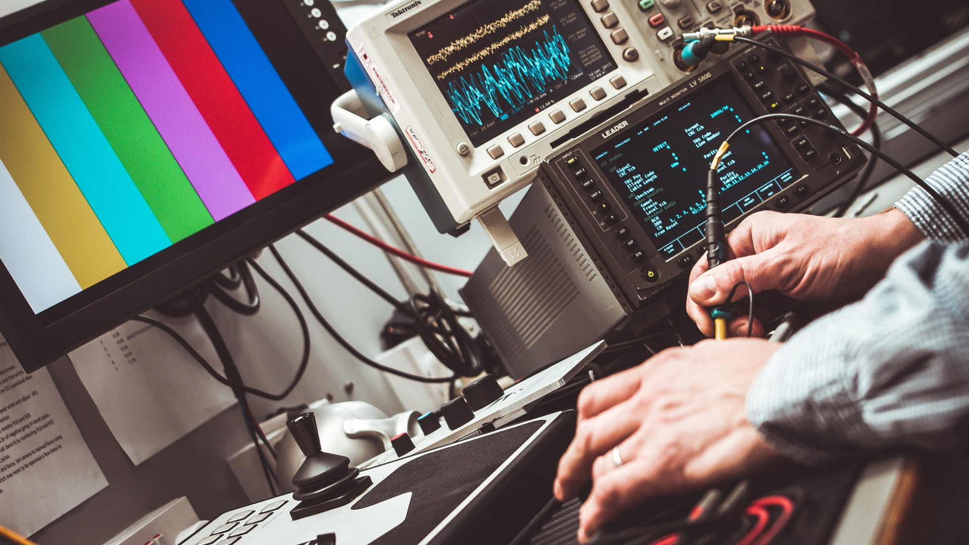 A monitor with color bars surrounded by measuring equipment operated by human hands with a stylus pen.