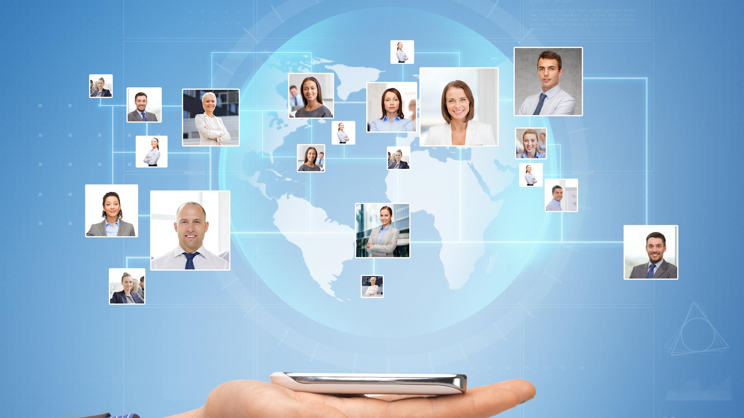 Hand holding smartphone with holographic display showing world map and floating portrait photos of diverse business professionals connected by network lines, representing global digital communication.