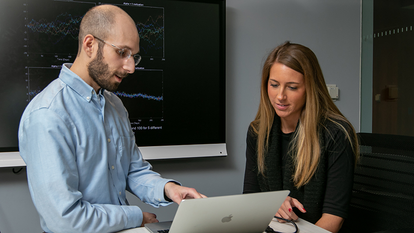 Professor Zachary Feinstein assists a student who is working on a laptop and there is a data chart in the background 