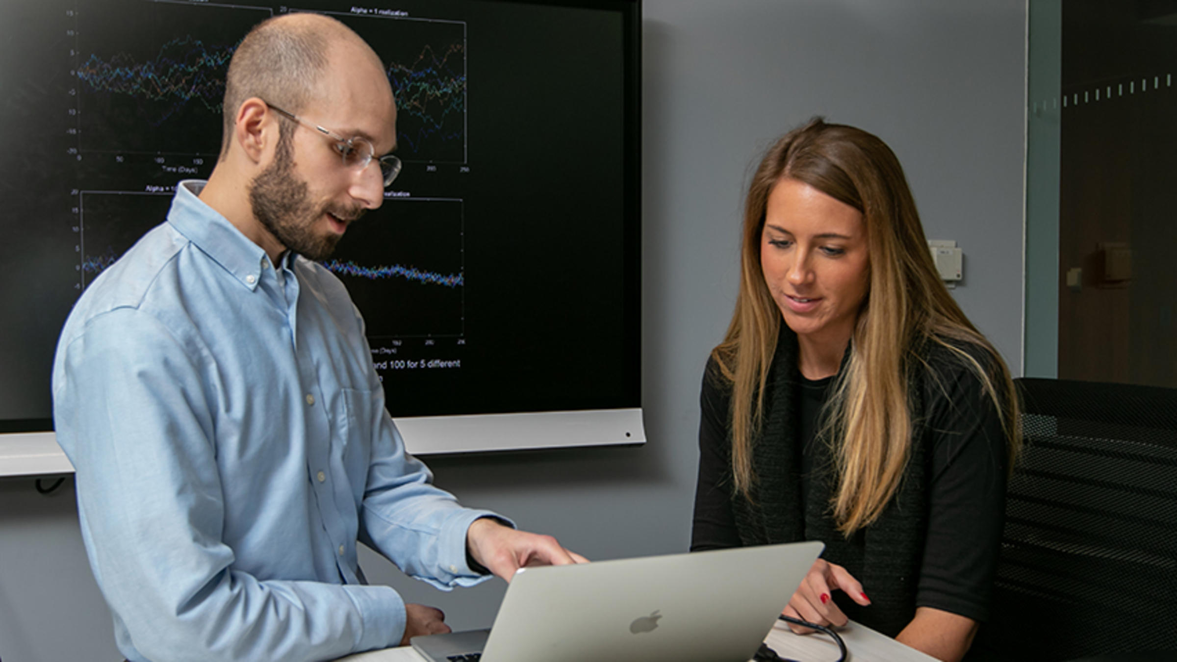 Professor Zachary Feinstein assists a student who is working on a laptop and there is a data chart in the background