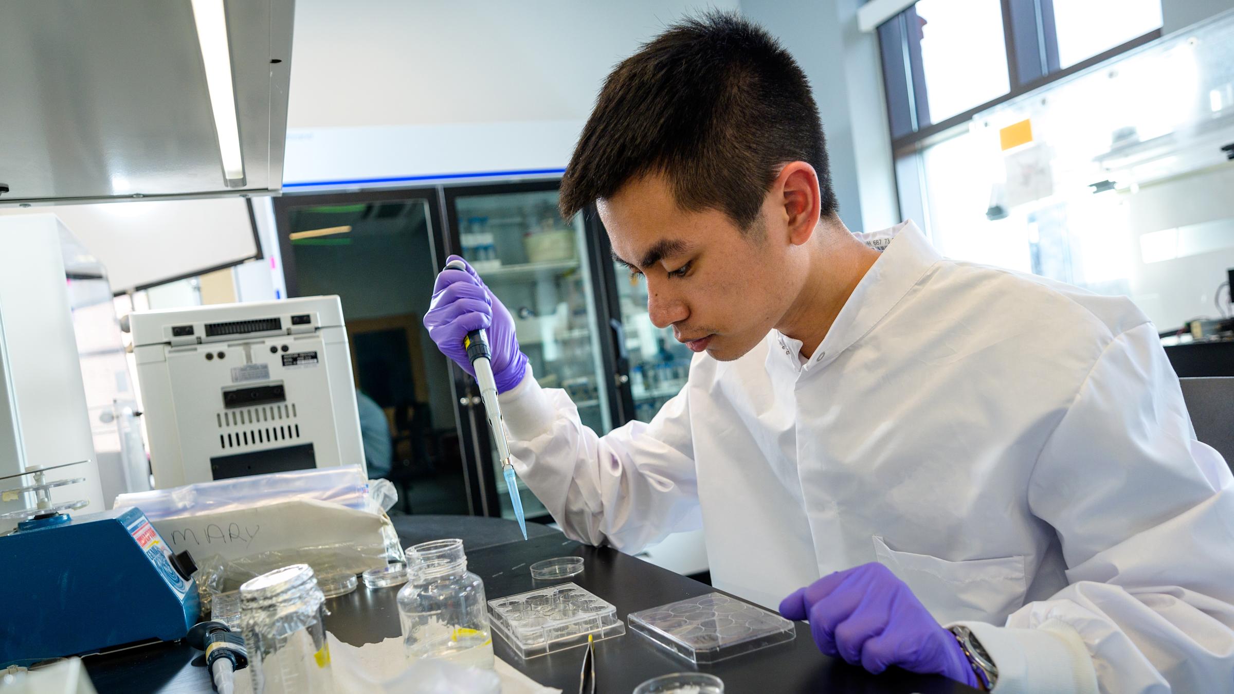 Student in lab coat uses dropper to view chemical sample.