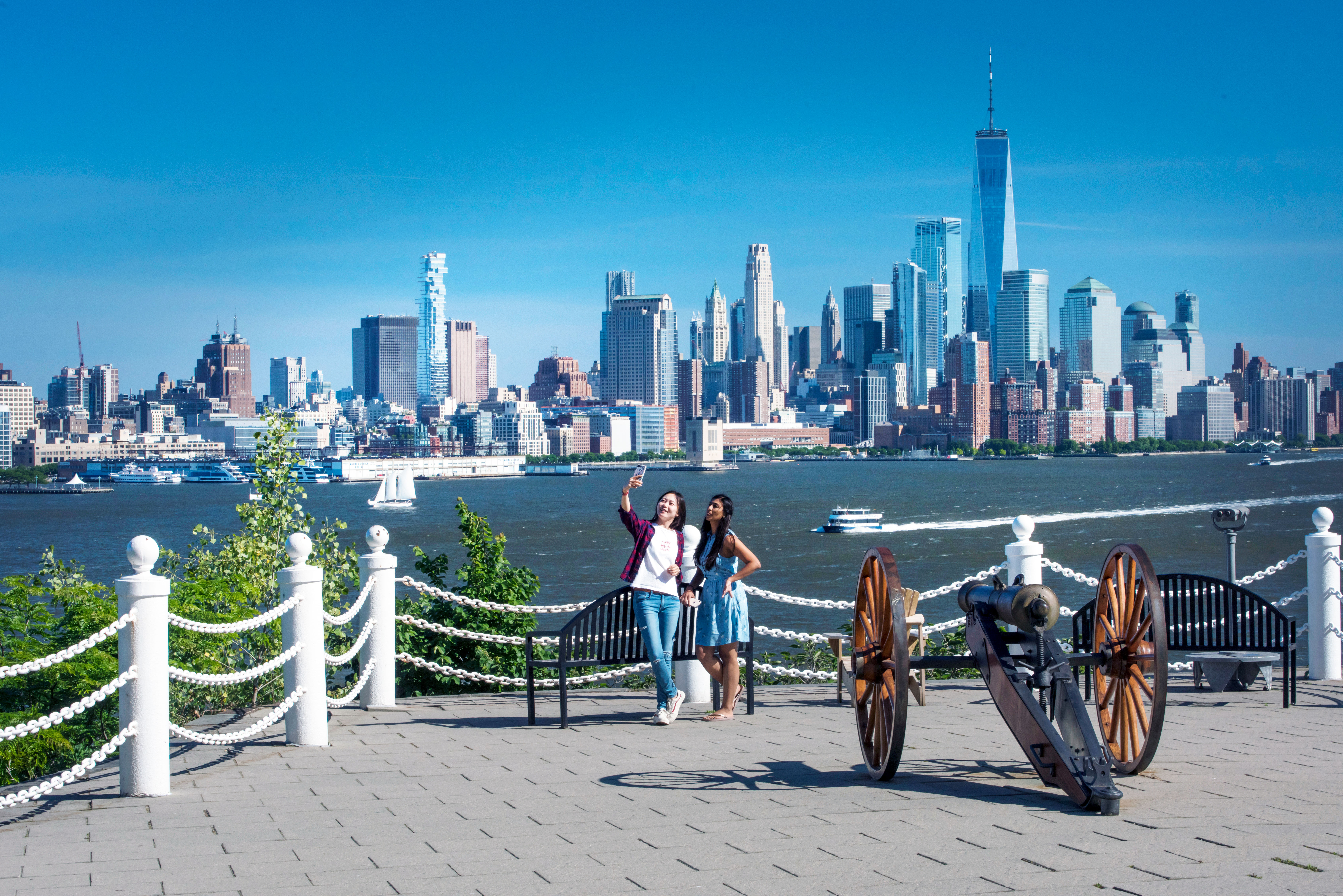 Two students take a photograph next to an antique cannon in Hoboken in front of the New York City skyline.