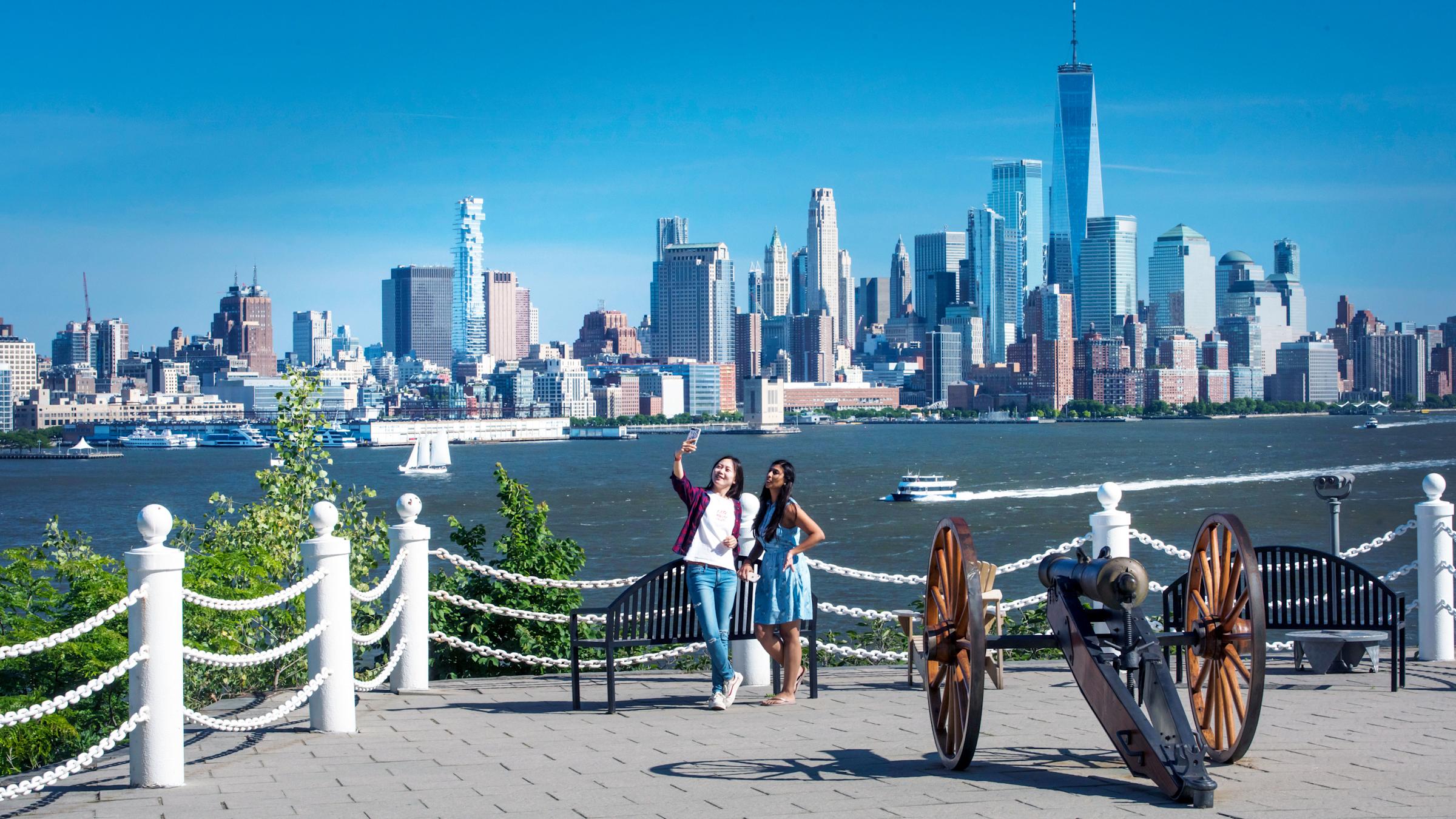 Two students take a photograph next to an antique cannon in Hoboken in front of the New York City skyline.
