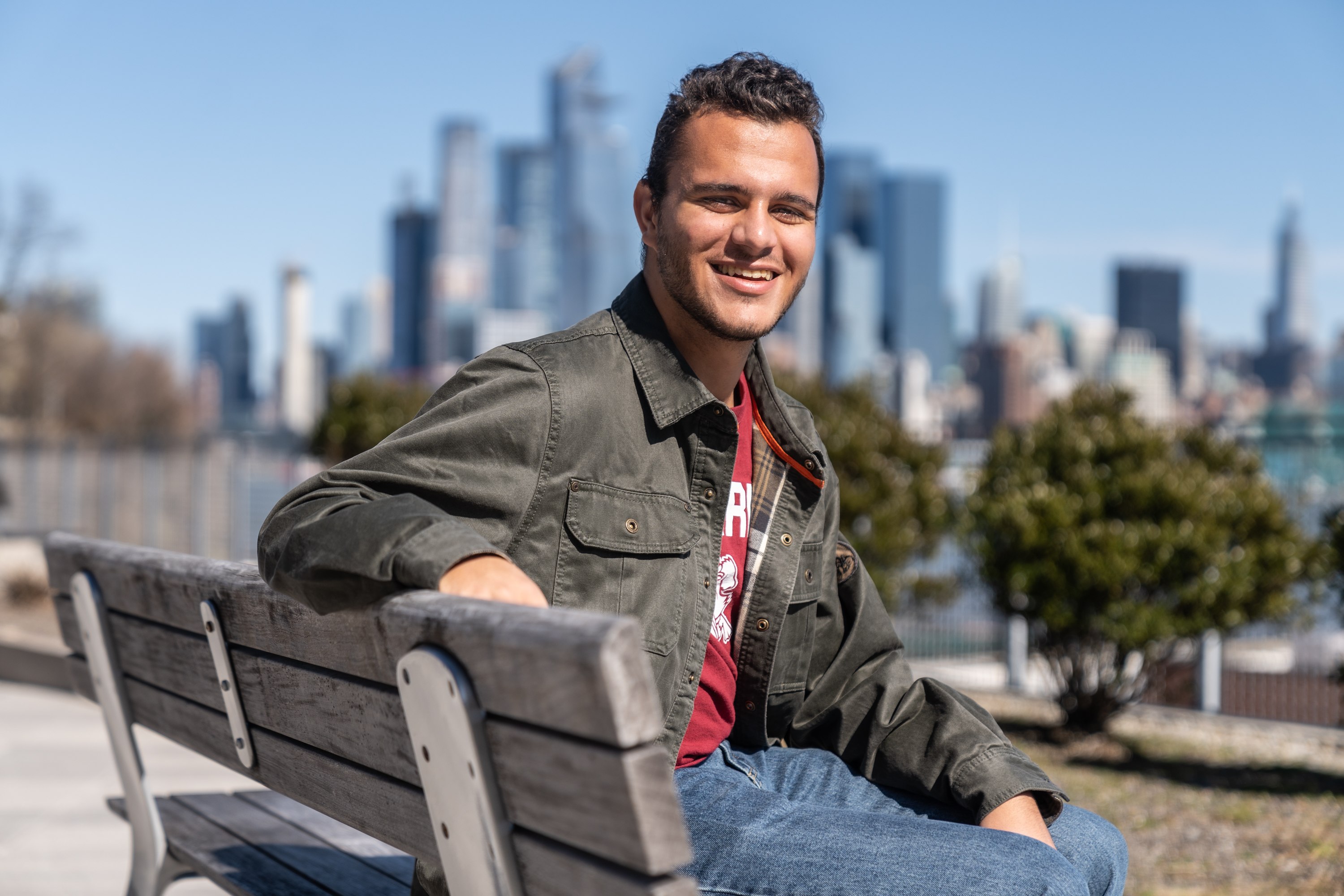 Gareth Maritz '23 sits on a bench with part of the New York City skyline in view behind him on a sunny day. 