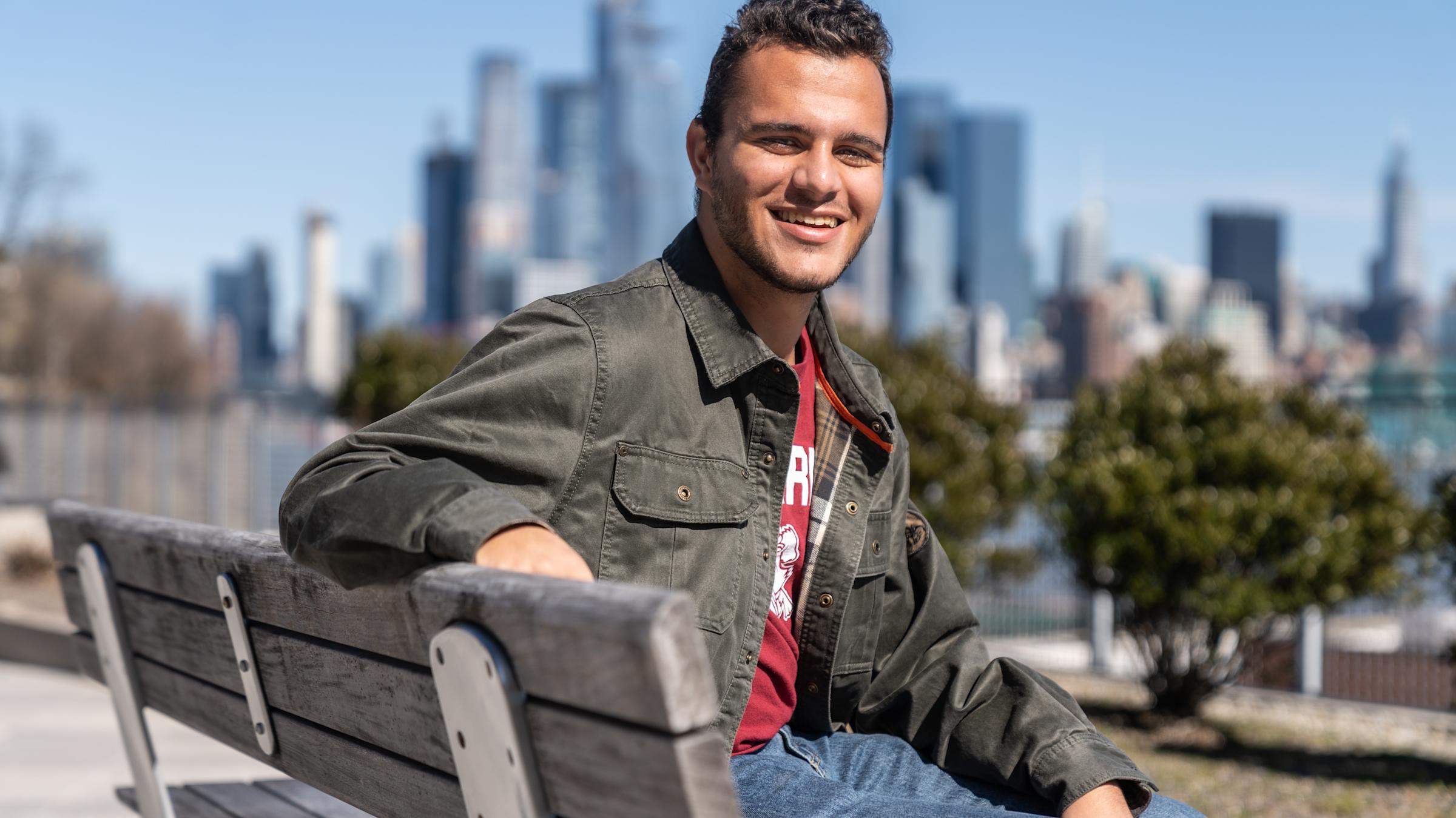 Gareth Maritz '23 sits on a bench with part of the New York City skyline in view behind him on a sunny day.